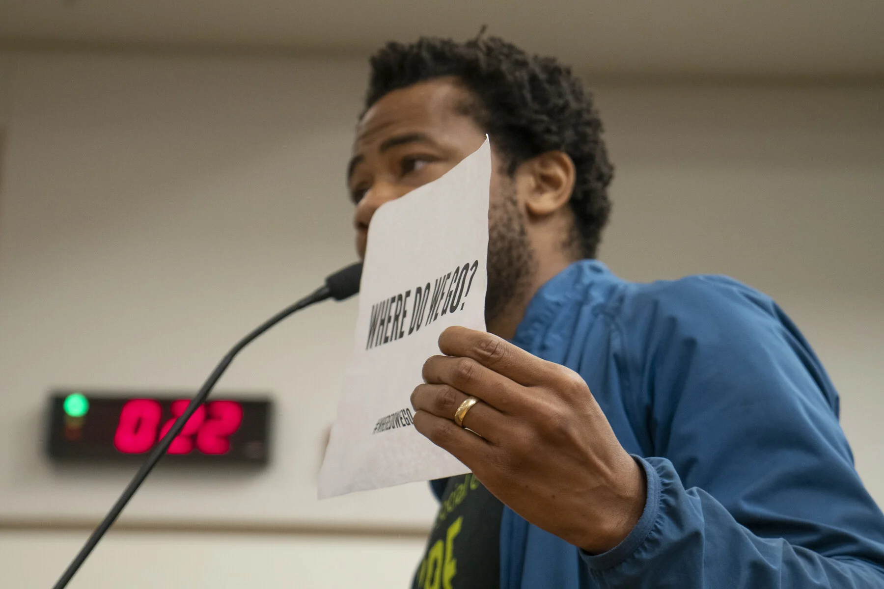 Aidan Hill speaking at a public meeting while holding a paper that reads "Where do we go?"