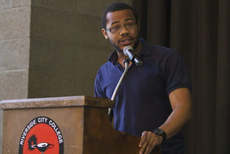 Aidan with glasses, wearing a blue shirt, speaking into a microphone at a Riverside City College podium.