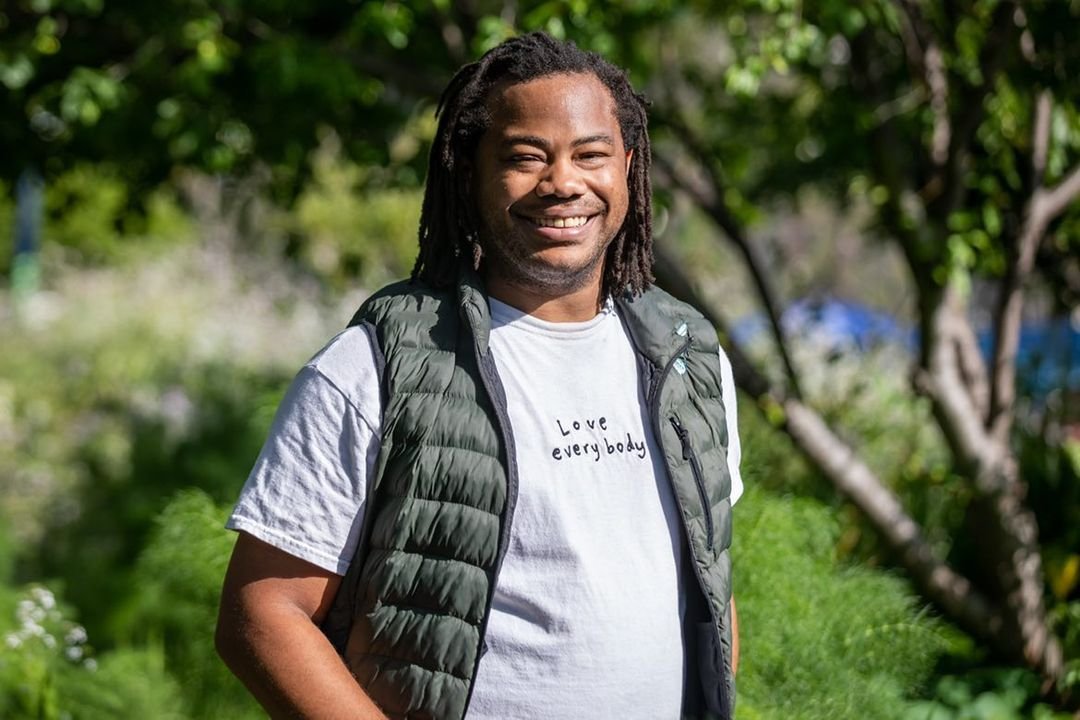 Aidan Hill smiling outdoors with trees in the background, wearing a white t-shirt and a black puffer vest.