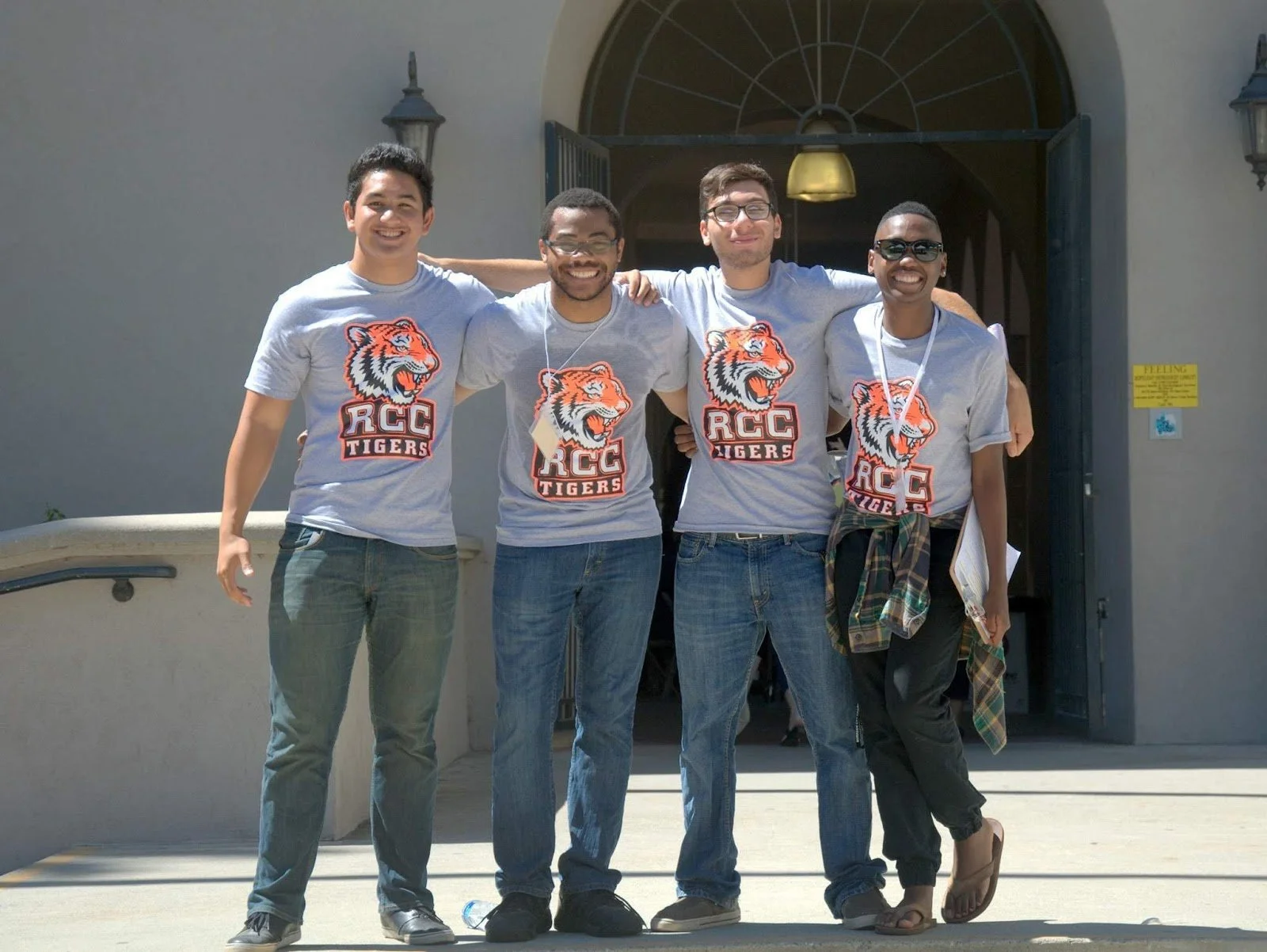 Four young adults including Aidan Hill standing together outside, smiling, wearing matching gray t-shirts with an orange tiger logo and the text 'RCC Tigers', with their arms around each other's shoulders.