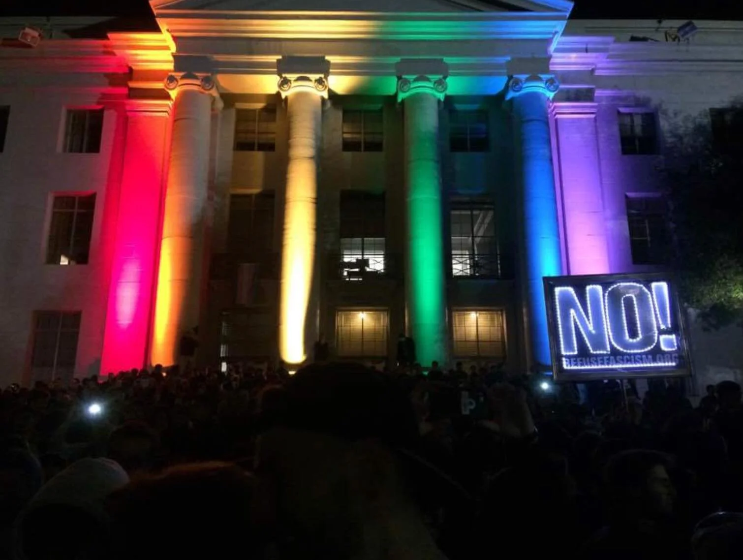 A crowd gathered outside UC Berkeley during a protest with Sproul Plaza illuminated with multicolored lights in rainbow order on columns, with a sign reading 'NO!'
