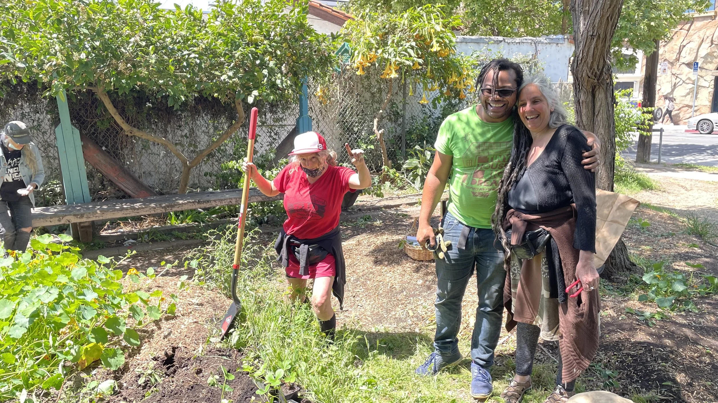 Group of four people in a garden, smiling and enjoying outdoor gardening activity on sunny day, with greenery and trees around.