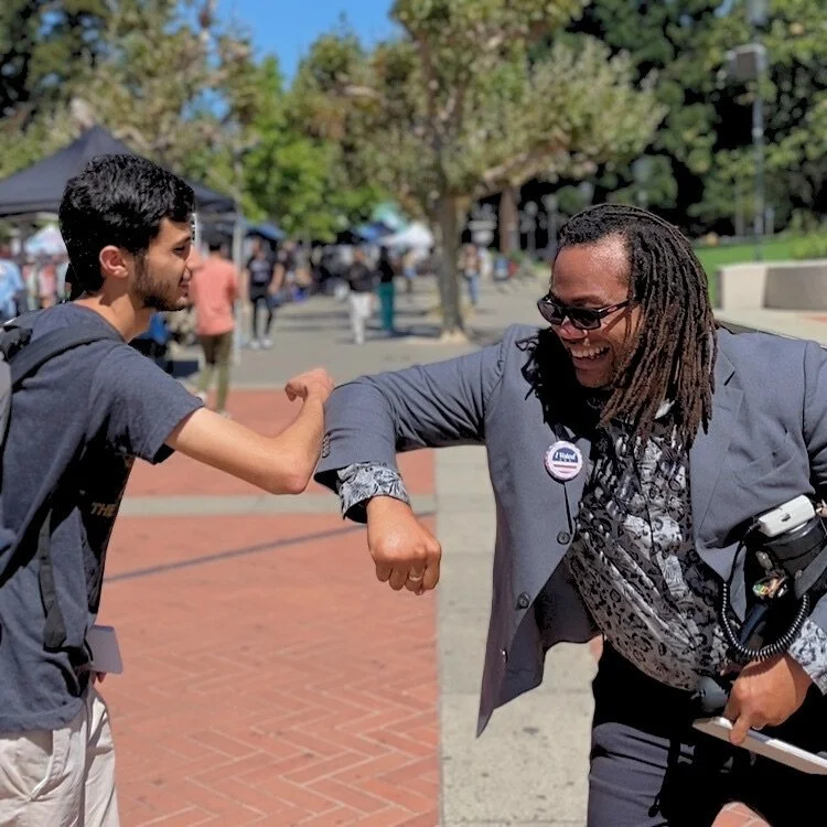 Aidan Hill and a UC Berkeley student greeting each other on Sproul Plaza with an elbow bump outdoors on a sunny day, with trees and people in the background.