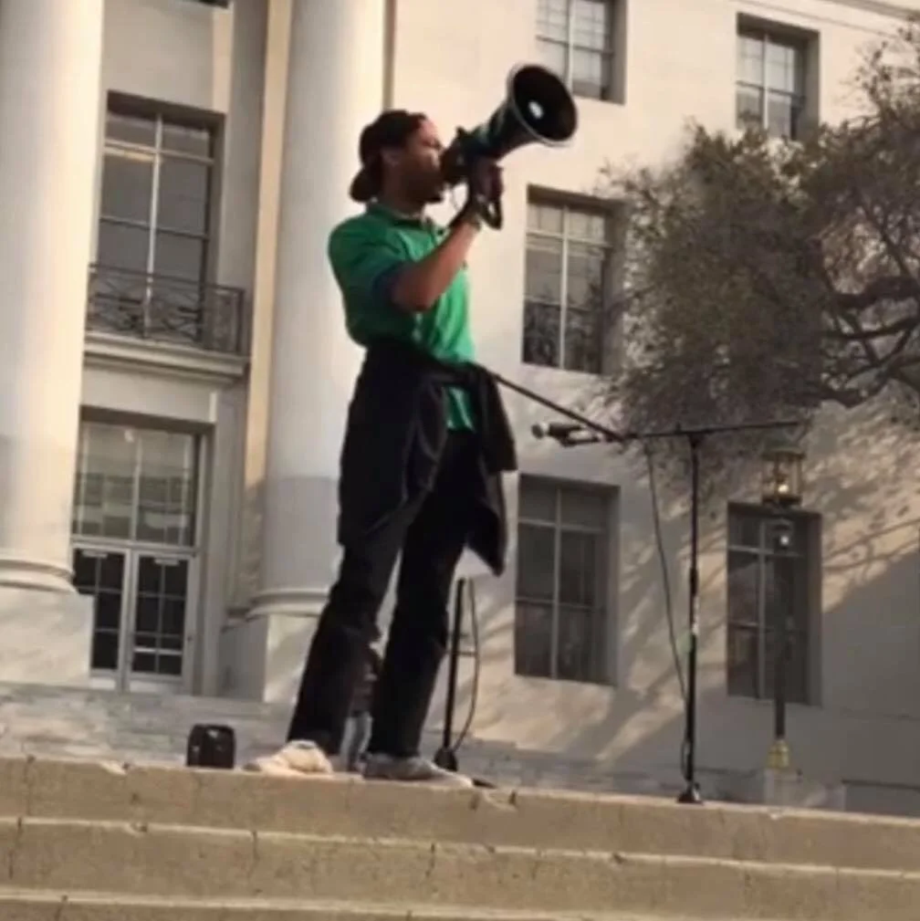 Aidan Hill standing outside on steps, speaking into a megaphone, wearing a green shirt with a black jacket tied around their waist. In the background, a building with large windows and trees are visible.