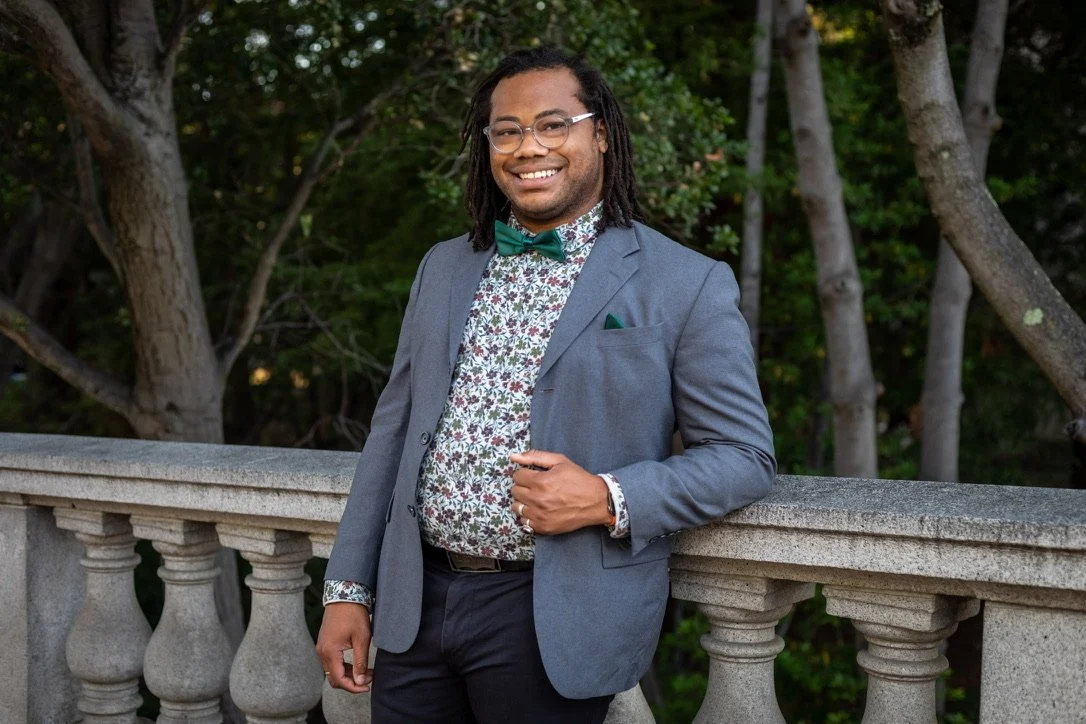 Aidan Hill standing outdoors by a stone railing in a gray suit jacket, floral shirt, green bow tie, and locs.