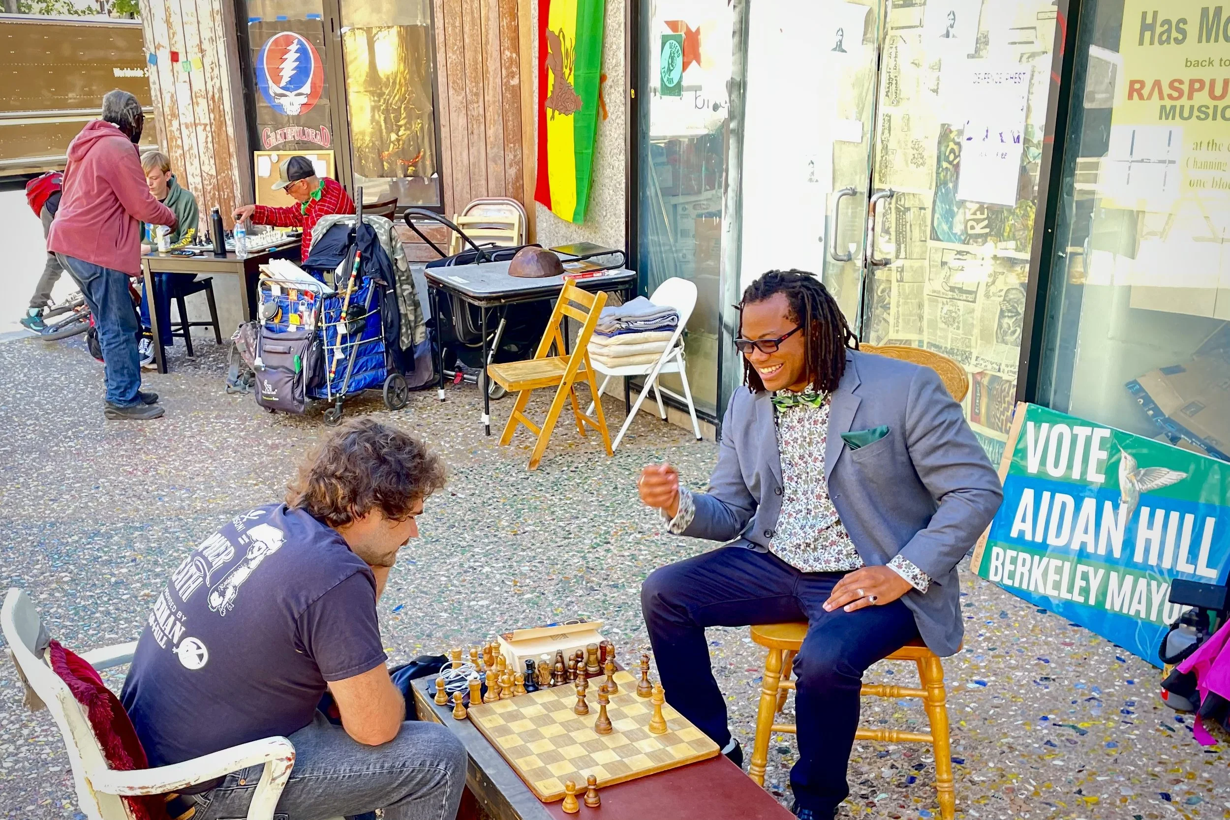 Two people playing chess on a city sidewalk, with Aidan Hill smiling and dressed in a suit, and the other wearing casual clothes. In the background, people are gathered around tables, and there is a sign supporting Aidan Hill for Berkeley mayor.