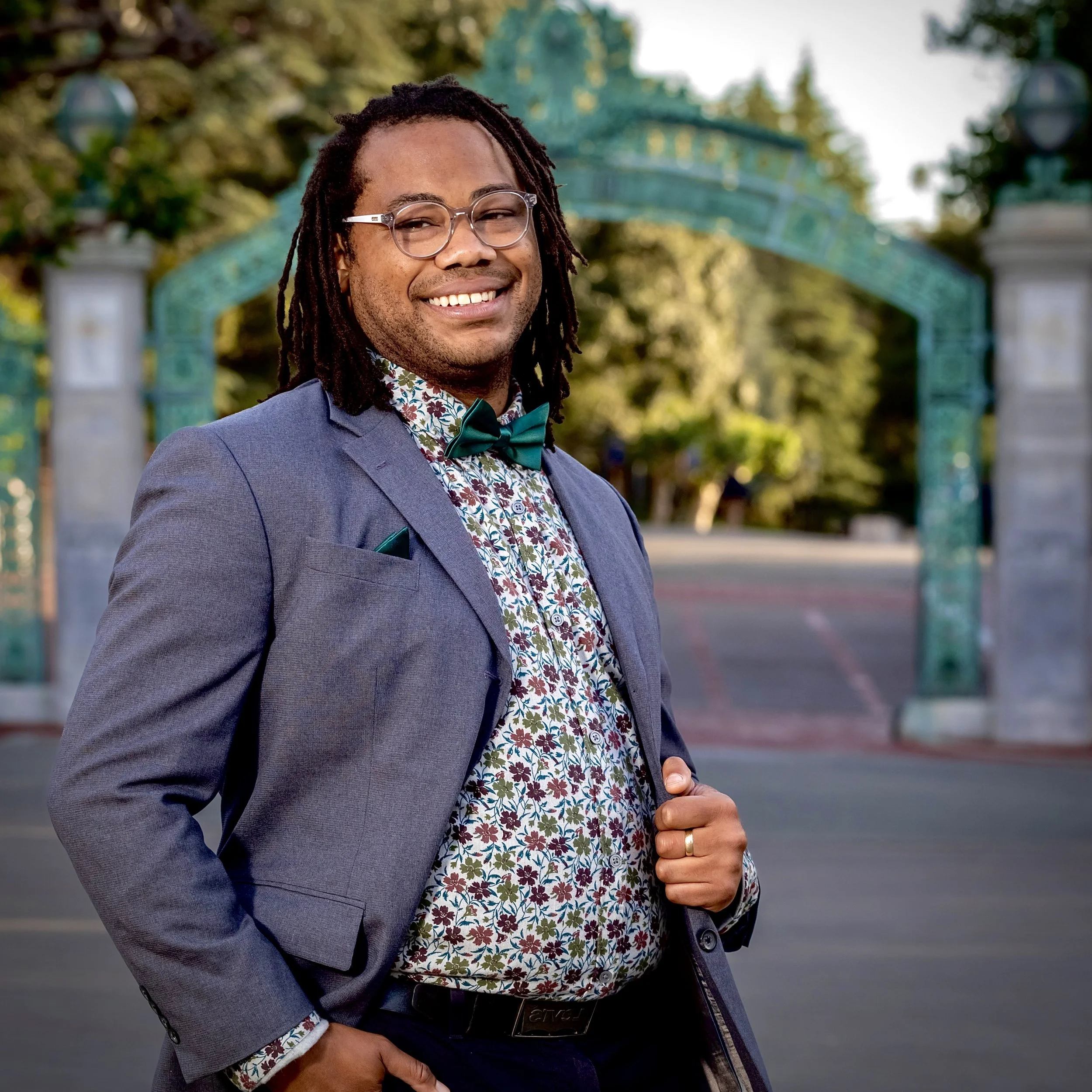 Aidan Hill smiling at Sather Gate in a gray suit jacket, floral shirt, green bow tie, and locs.
