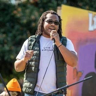 Aidan Hill with locs, sunglasses, and a microphone speaking at an outdoor event, wearing a white t-shirt and a black vest, with a colorful background.