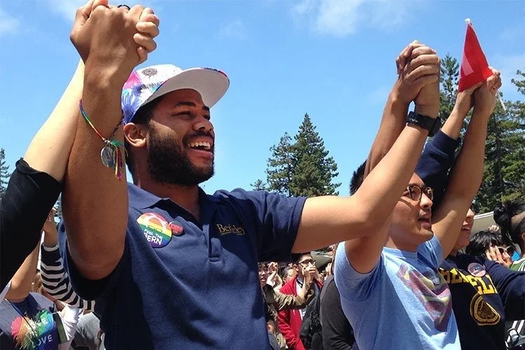 Aidan Hill holding hands with people at a pride parade, raising their arms in celebration, with trees and a blue sky in the background.