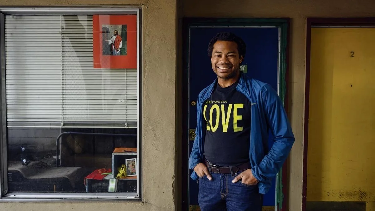 Aidan Hill smiling with dark hair and medium skin tone wearing a blue jacket and a black T-shirt with yellow text that says 'LOVE', standing in front of a blue door, with a window showing an interior space with blinds and a picture of Jesus Christ.