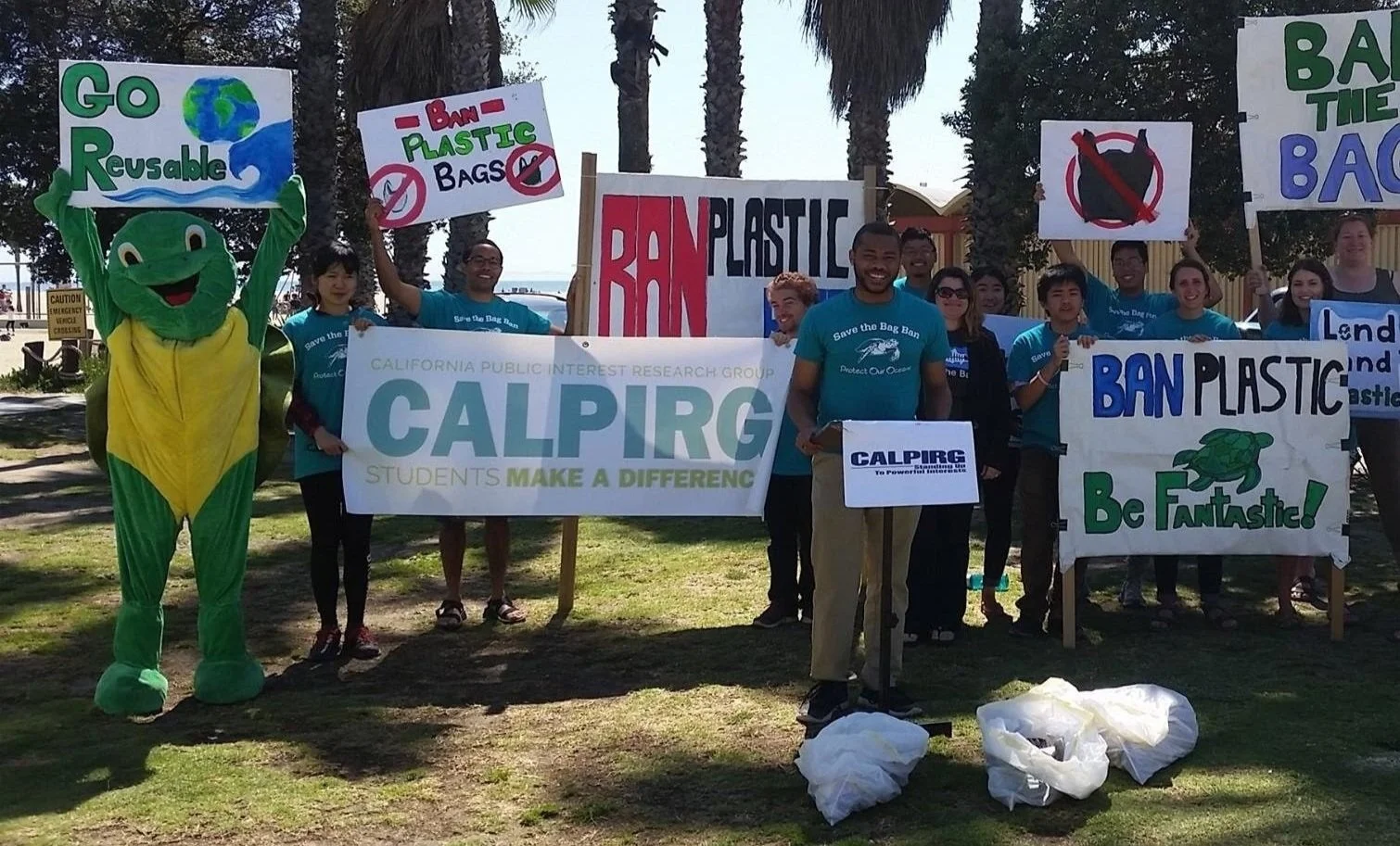 Group of people protesting against plastic bags, holding signs that say 'Ban Plastic Bags,' 'Go Reusable,' and 'Ban Plastic, Be Fantastic!' One person is dressed in a turtle costume, and another holds a large banner that reads 'California Public Interest Research Group - Students Make a Difference.'