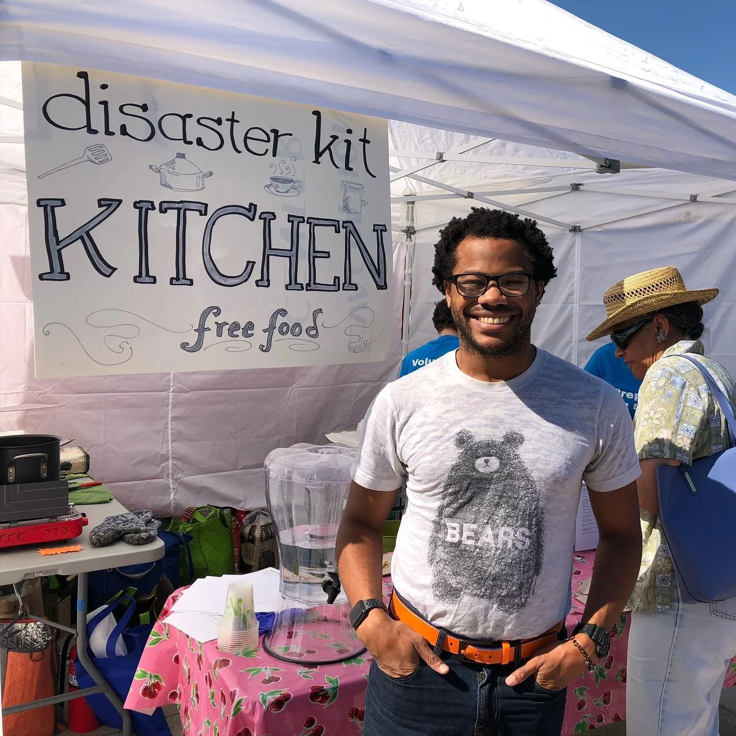 Aidan Hill smiling standing in front of a table with a sign that reads "disaster kit kitchen free food" at an outdoor event. The table has a water pitcher and other items. Other people are in the background under the tent.