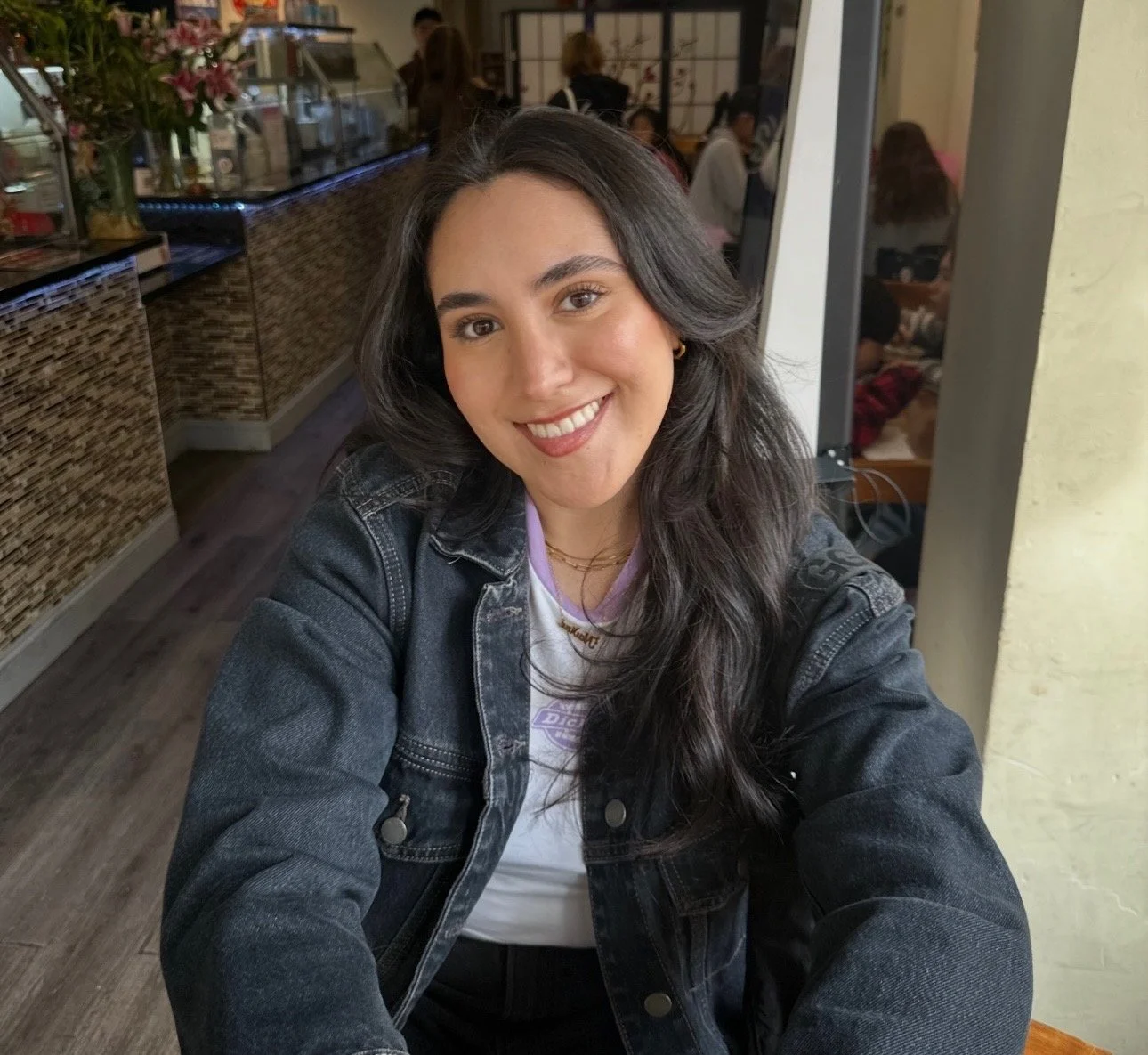 A young woman with long dark hair, smiling, wearing a denim jacket and white shirt sitting in a restaurant or cafe.
