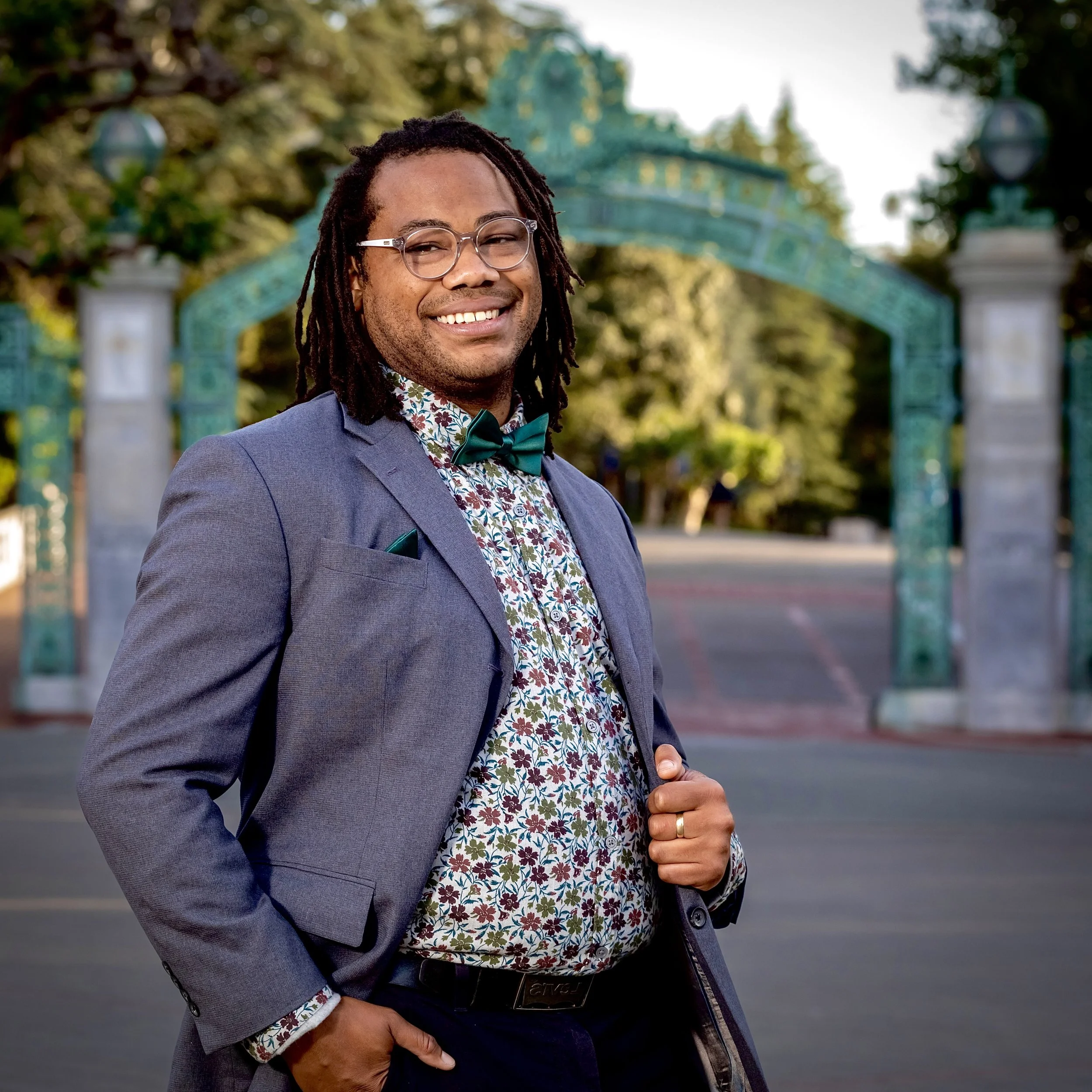 Aidan Hill smiling with glasses and locs, in a suit with a floral shirt and bow tie, standing outdoors near Sather Gate at UC Berkeley, an ornate green archway.