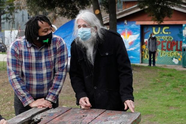 Aidan Hill and Michael Delacour wearing face masks and jackets talking outdoors in front of a graffiti-covered building at People's Park with a person standing in the background.
