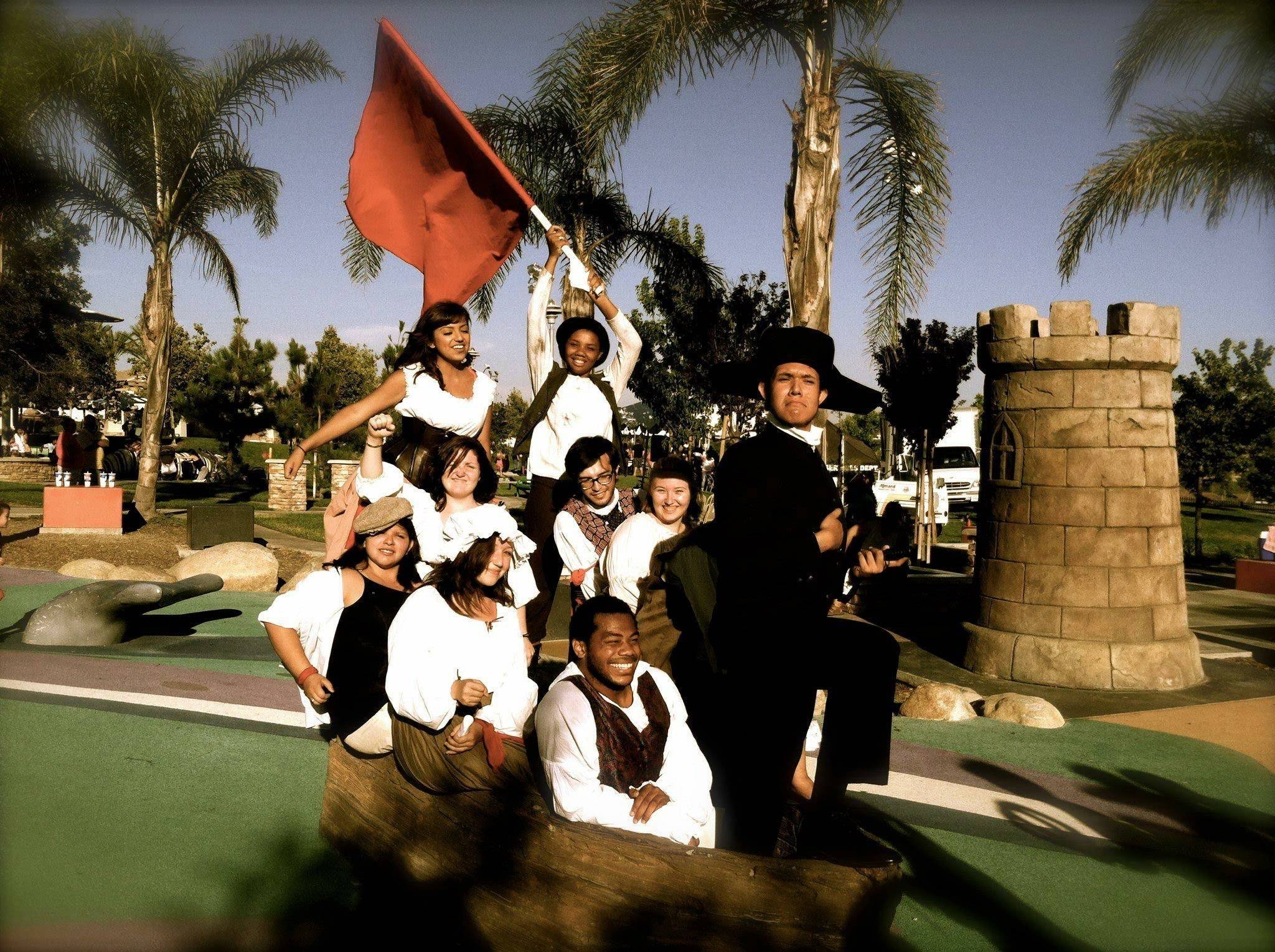 Aidan Hill and a group of people dressed in colonial costumes pose together outdoors at a park with palm trees. They appear to be participating in a themed event, with some sitting and others standing, holding a flag, and wearing historical attire.
