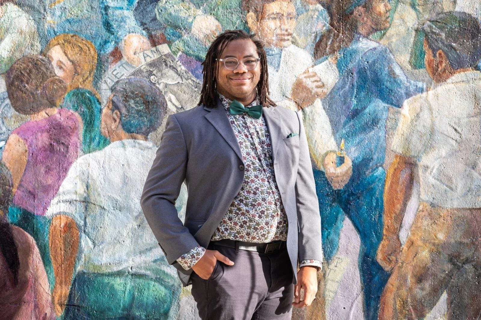Aidan Hill in a gray suit, floral shirt, and green bow tie standing in front of a colorful mural of people.
