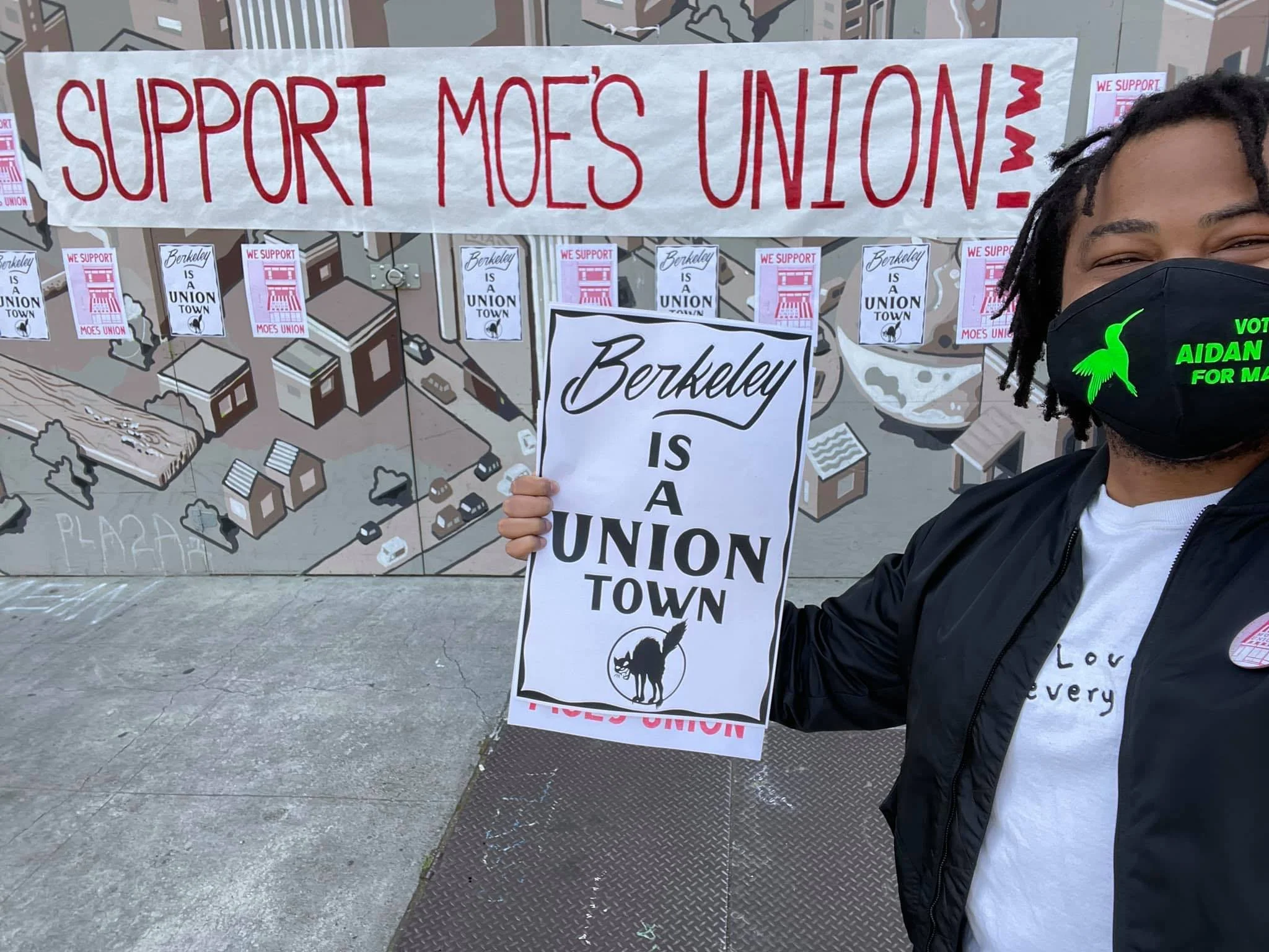 Aidan Hill wearing a black mask with a green hummingbird logo and the words 'VOTE AIDAN FOR MAYOR' holding a sign that reads 'Berkeley is a union town' in front of a mural and protest signs that say 'Support Moes Union'