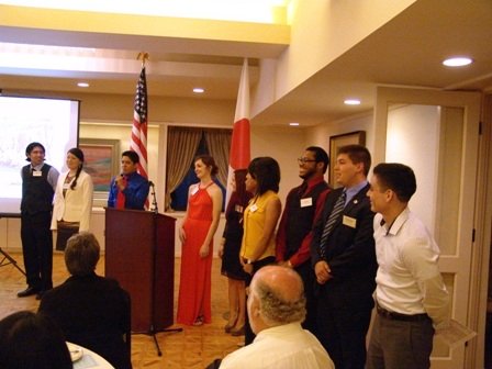 Aidan Hill among a group of people standing in a line during a formal event, with flags and an audience seated in the room.