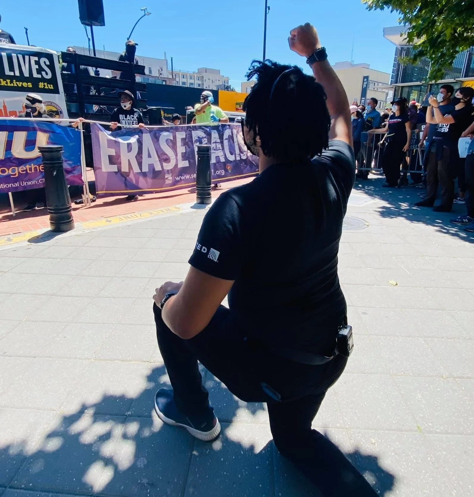 Aidan Hill kneeling on one knee with their fist raised in front of a crowd at a protest or rally. The crowd is behind a barrier with signs, and other protesters are visible, many wearing masks.