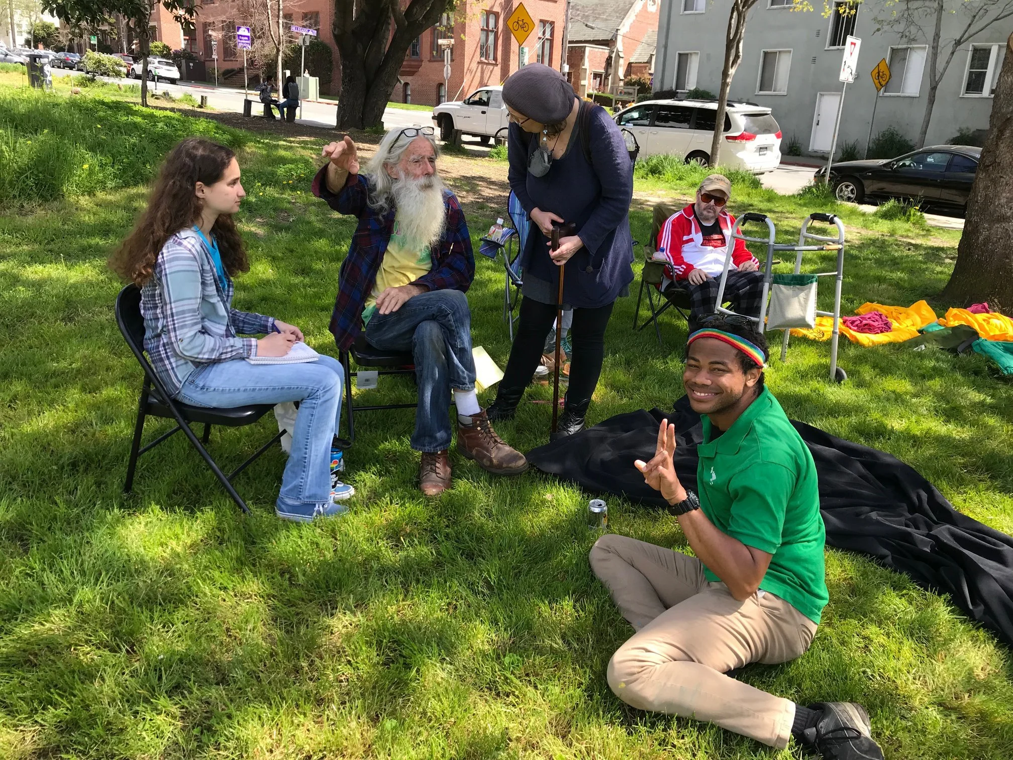 Aidan Hill among a group of people having an outdoor discussion in a park, sitting on chairs and the grass, with trees and buildings in the background.