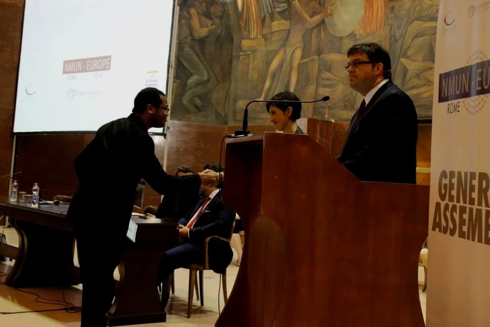 Aidan Hill in a suit shaking hands with another man in a suit at a conference, with a woman in the background near a podium and others seated at a table, in a room with a mural and conference banners.
