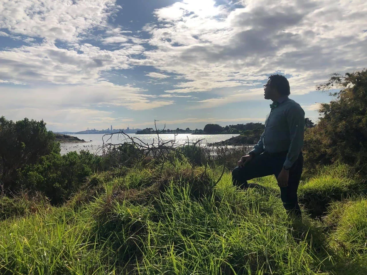 Aidan Hill in a long-sleeved shirt and glasses sitting on grass near a body of water with a city skyline in the background, under a cloudy sky.