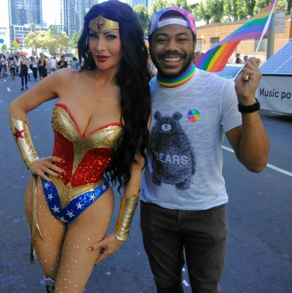 A woman dressed as Wonder Woman standing next to a Aidan Hill wearing rainbow-themed LGBTQ+ pride accessories at a pride parade.