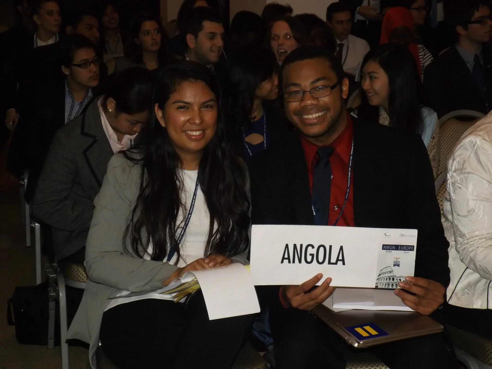 A group of people attending an international conference, with Aidan Hill and a young woman holding a sign that reads 'Angola'.