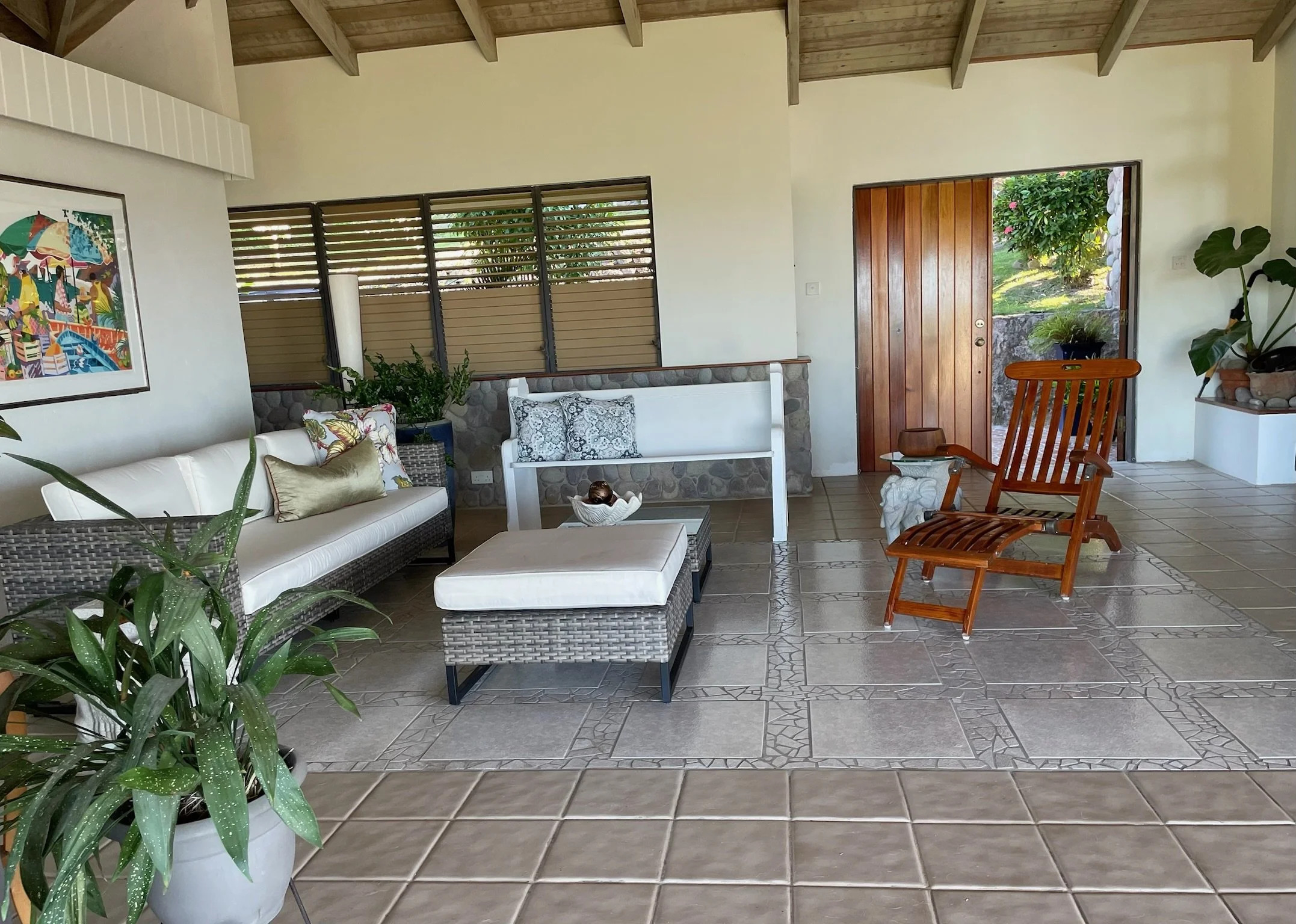Living room with a white sofa, a matching ottoman, a wooden recliner chair, and a white bench, with potted plants, colorful pillows, and a wooden door leading outside with greenery visible.