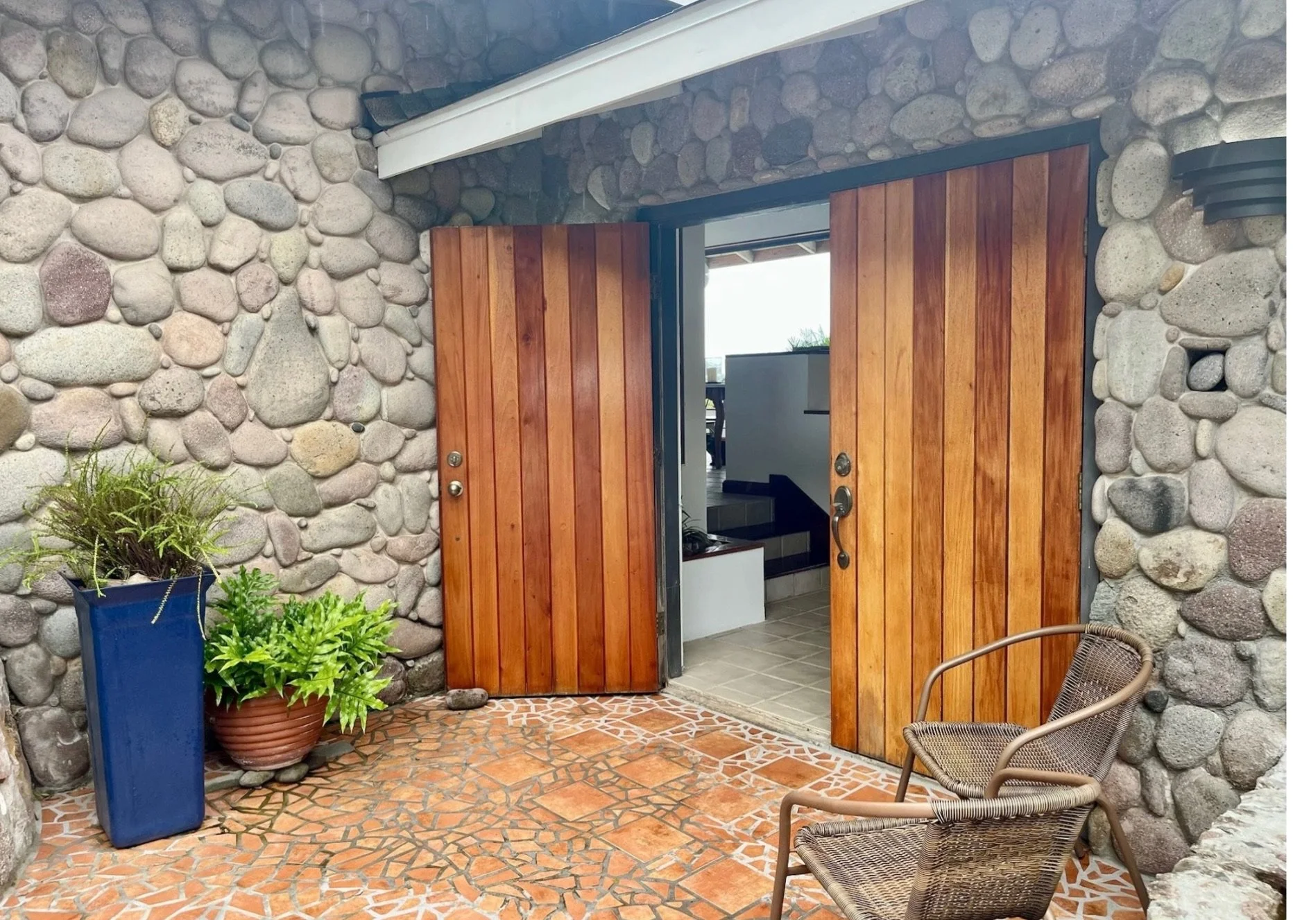 A stone wall entryway with double wooden doors open to an indoor area with stairs and tiled floor, decorated with potted plants and a wicker chair.