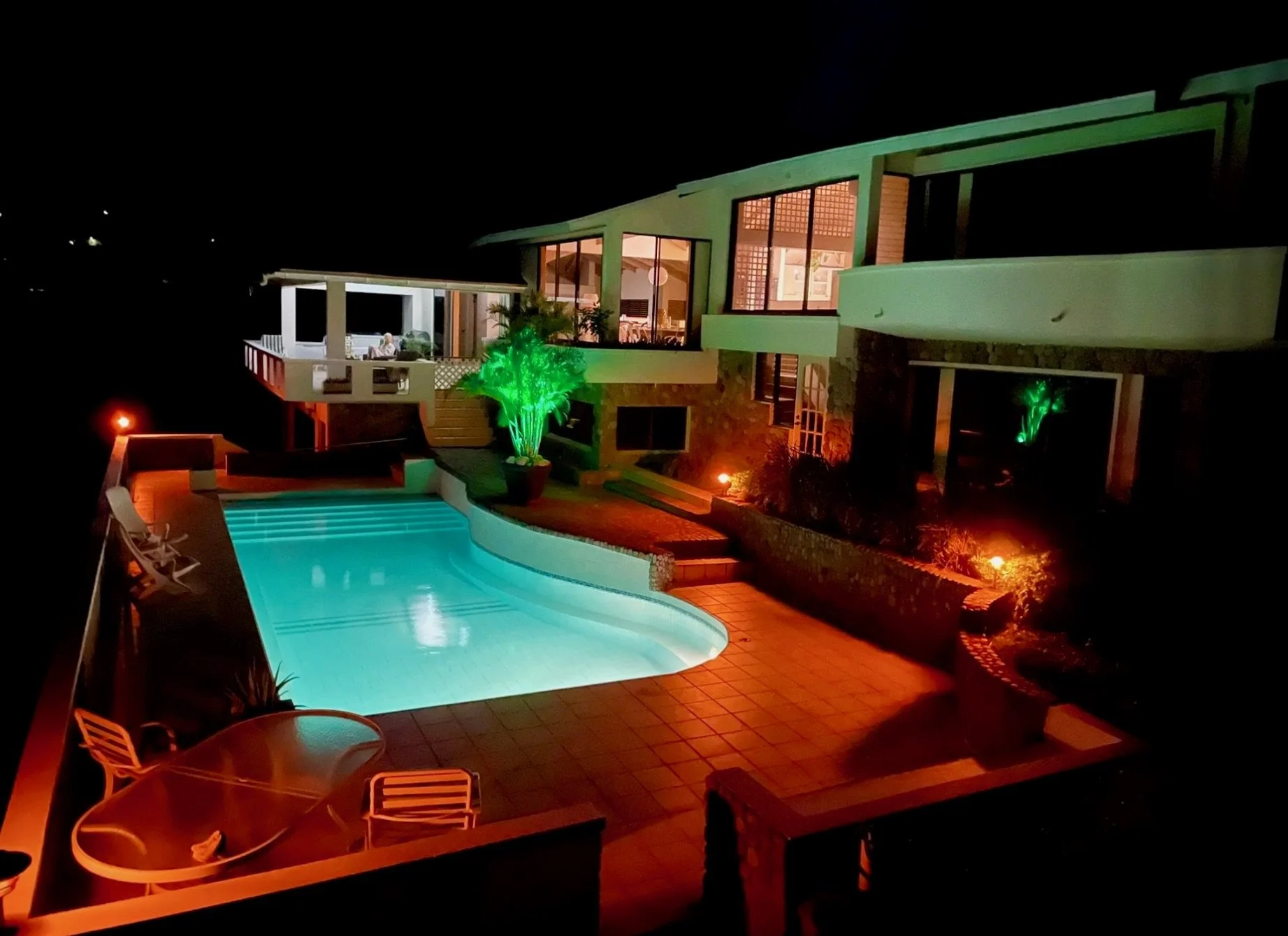 Night view of a modern house with illuminated swimming pool, outdoor seating, green-lit plants, and large windows showing the interior.