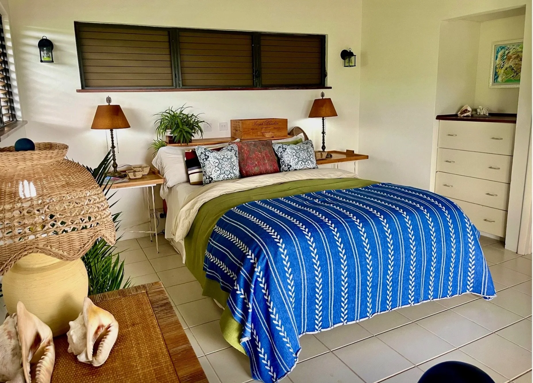 A cozy bedroom with a made bed covered in a blue and white patterned blanket and decorative pillows. There are wooden nightstands with lamps, a plant, and a window with wooden blinds on the wall behind the bed. A white dresser with decorative items i