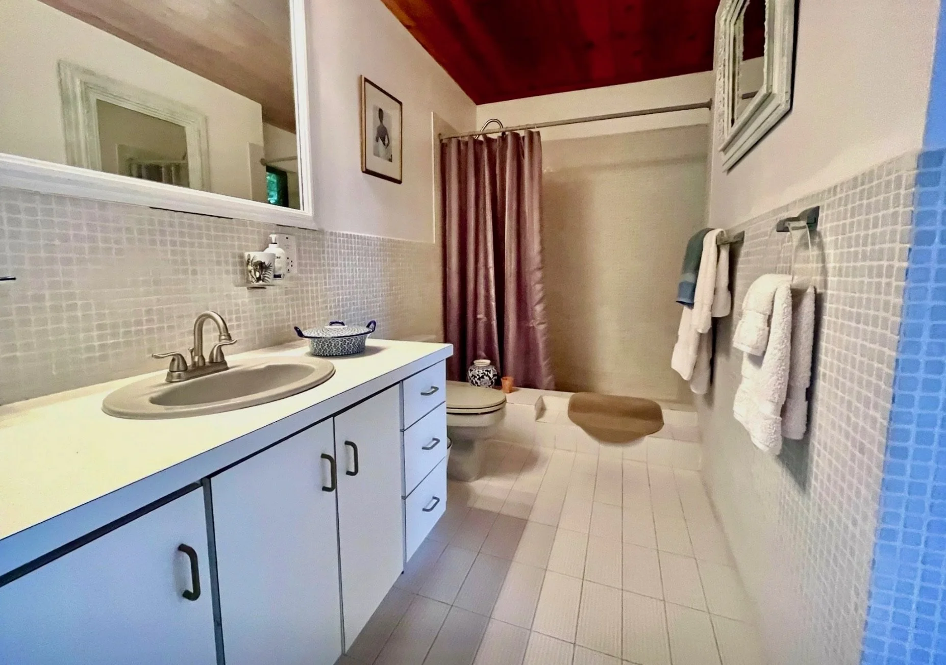 A bathroom with a white vanity and sink, a mirror above, a patterned basket, a toilet, and a shower with pink curtains. Towel racks with towels, framed artwork on the wall, and wood-paneled ceiling.