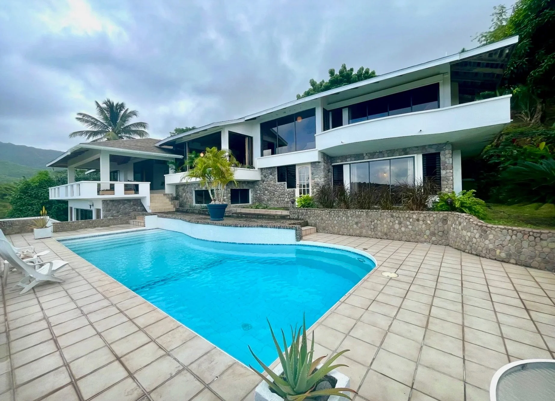 Modern house with large windows and stone walls, overlooking a swimming pool with blue water, surrounded by a tiled patio with lounge chairs and potted plants, with lush green trees and hills in the background under a cloudy sky.