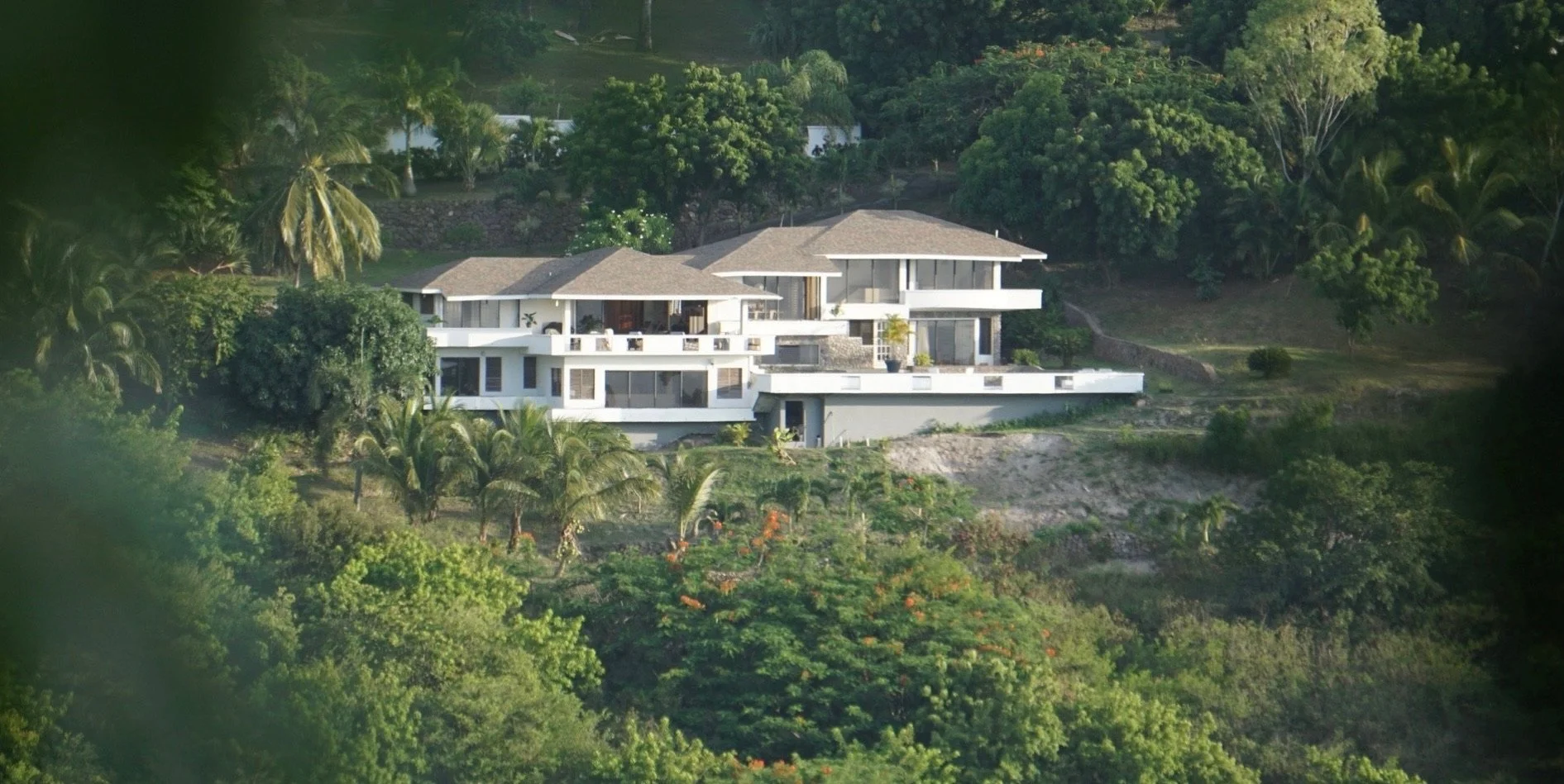 Modern white multi-level house on a hillside surrounded by lush green trees and vegetation.