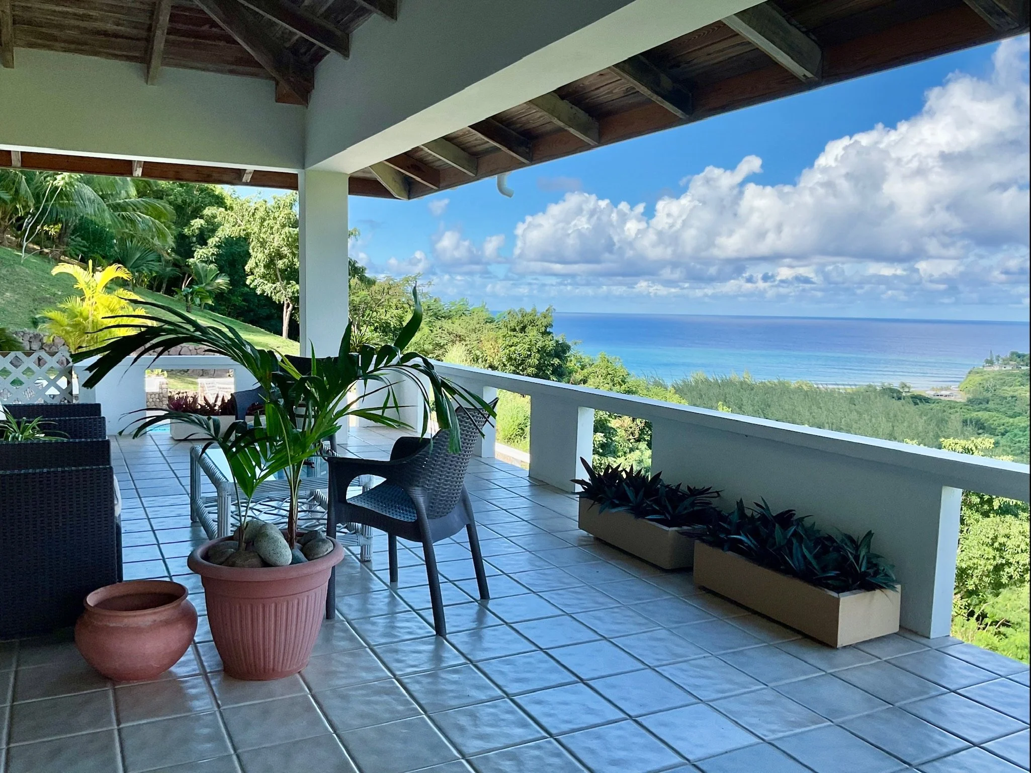 A spacious outdoor patio with tiled flooring, black wicker chairs, potted plants, and a view of green hills, water, and a partly cloudy sky in the distance.