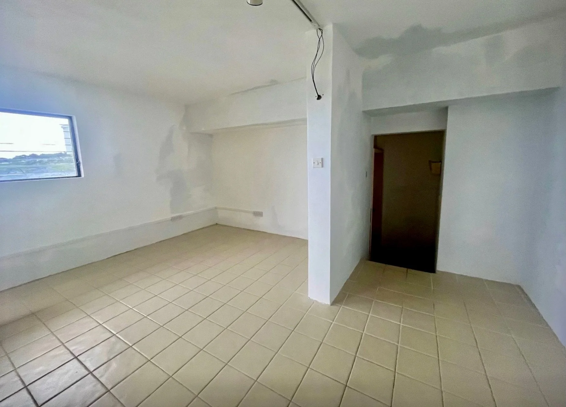 Interior of a room under renovation with beige tiled floor, white walls, and a small window on the left side. An unfinished ceiling with exposed wiring hangs from one corner.