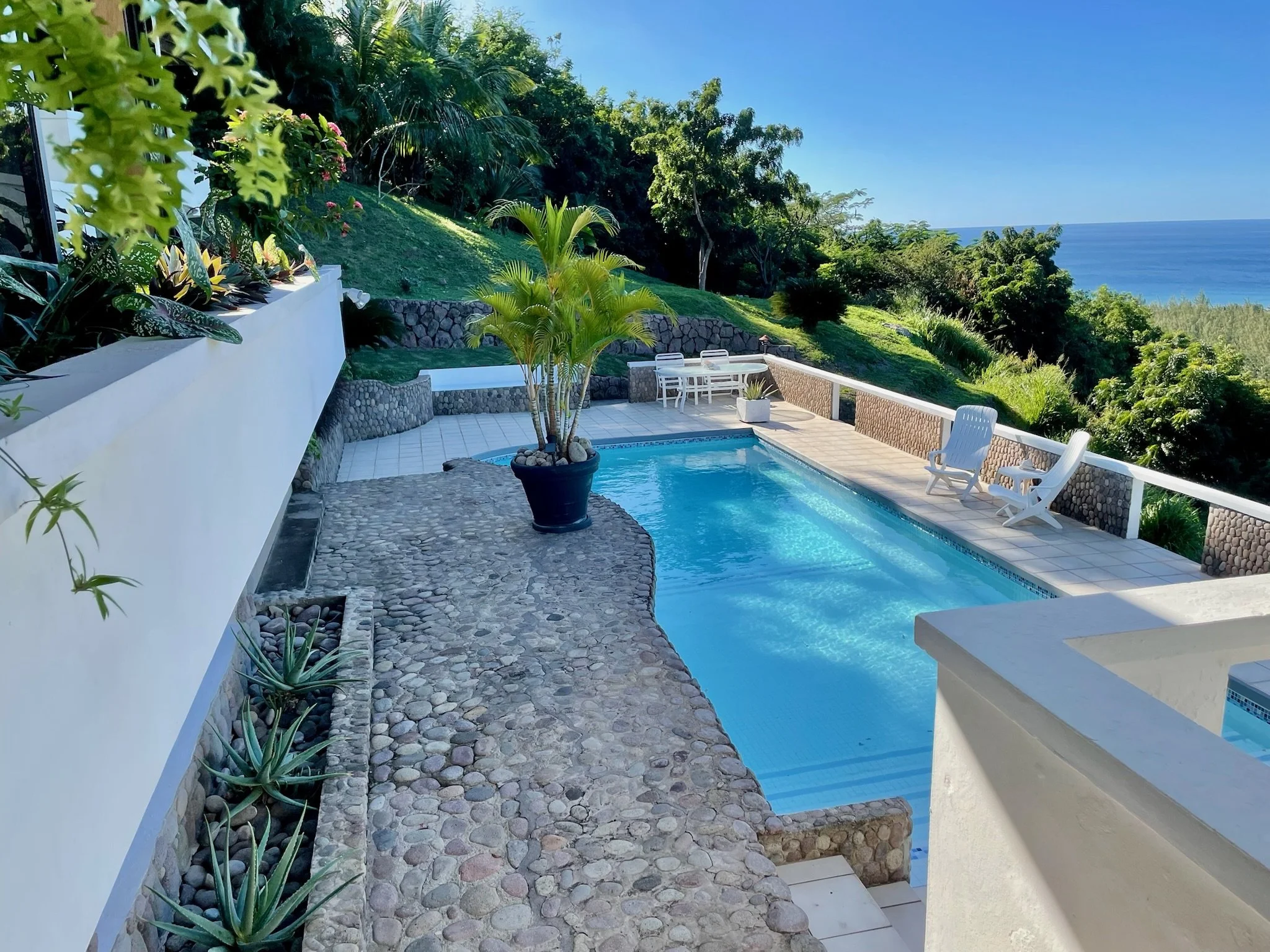 A backyard with a swimming pool overlooking the ocean, surrounded by greenery and a stone patio with white Adirondack chairs and a round table.