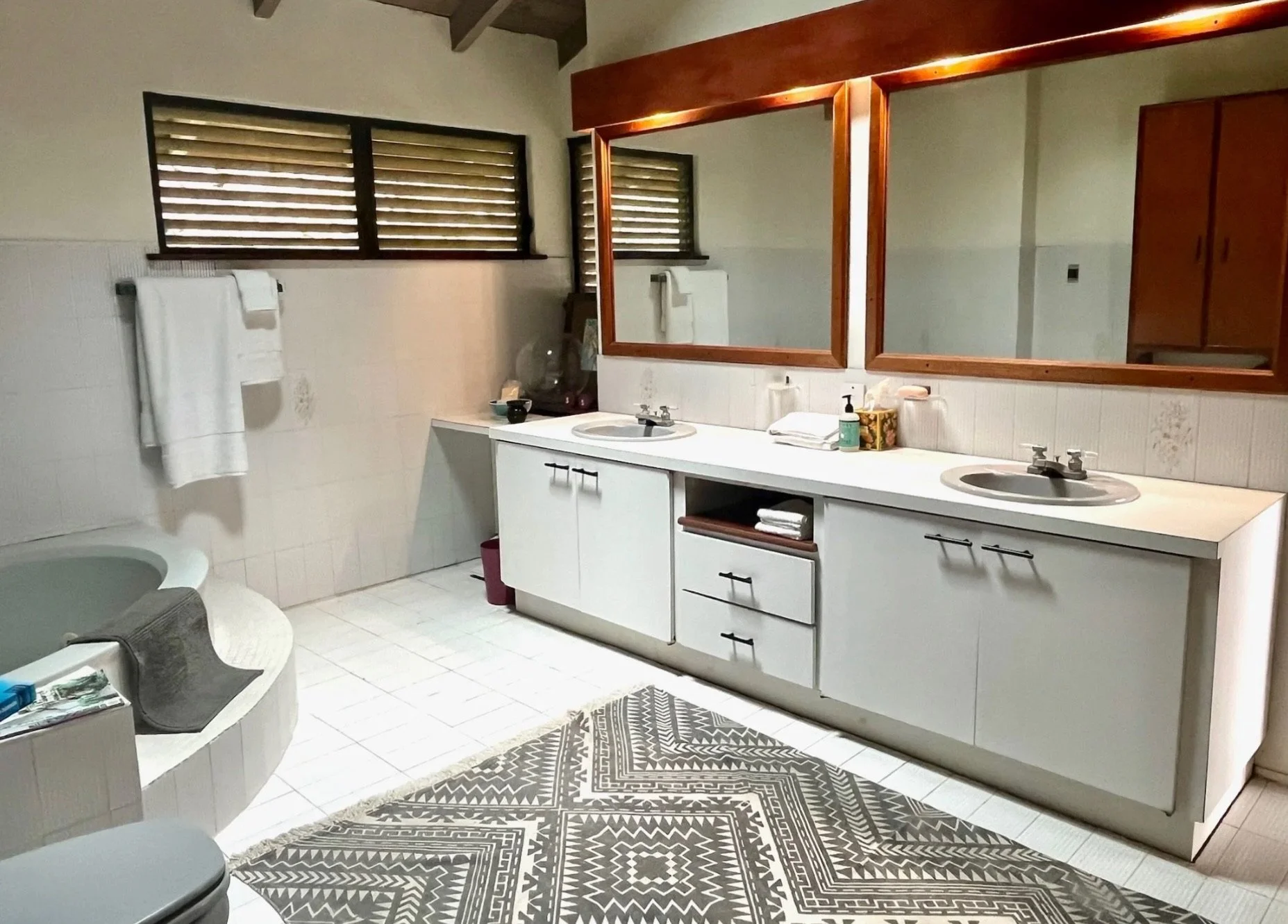 Bathroom with dual sinks, large wooden framed mirrors, white cabinets, and a bathtub in the corner with towels and toiletries.
