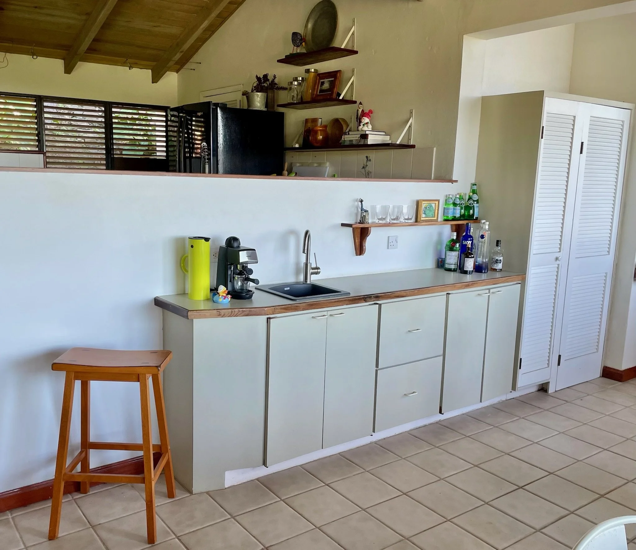 Kitchen counter with a coffee machine, a yellow thermos, and a small rubber duck. Behind the counter are cabinets and a shelf with glasses, bottles, and decorative items. To the right is a closed white louvered closet door. The floor is tiled, and th