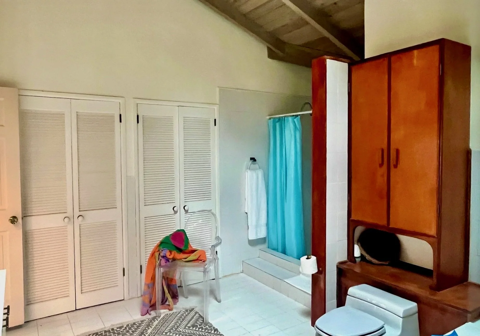 Interior view of a bedroom with closet doors, a chair with colorful towels, a staircase with a blue curtain, a towel hanging on a hook, and a wooden storage unit next to a toilet.