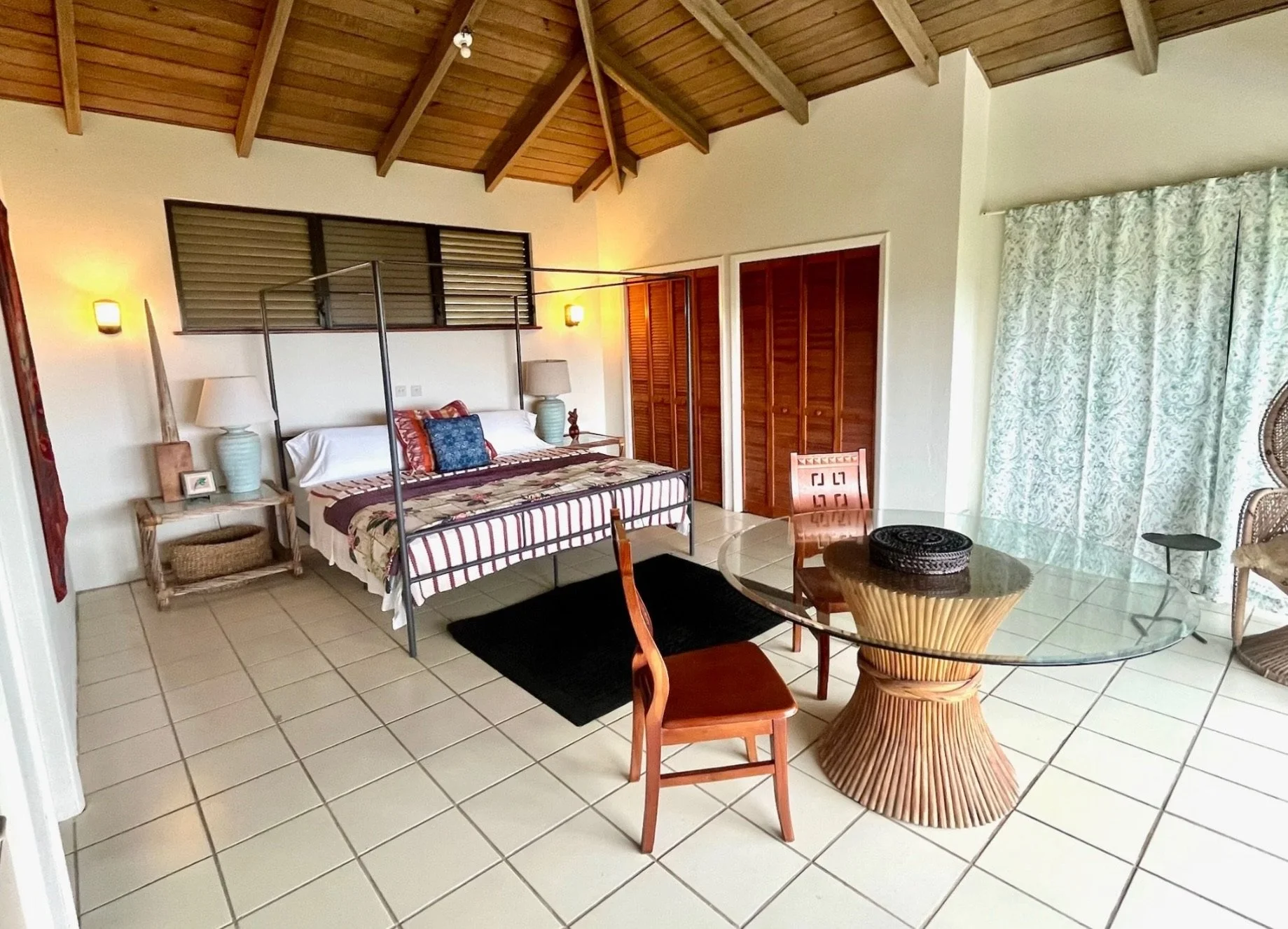 A tropical-style bedroom with a sloped wooden ceiling, a glass-top round dining table with wooden chairs, a bed with a colorful striped and floral bedspread, and large patterned curtains covering a window.