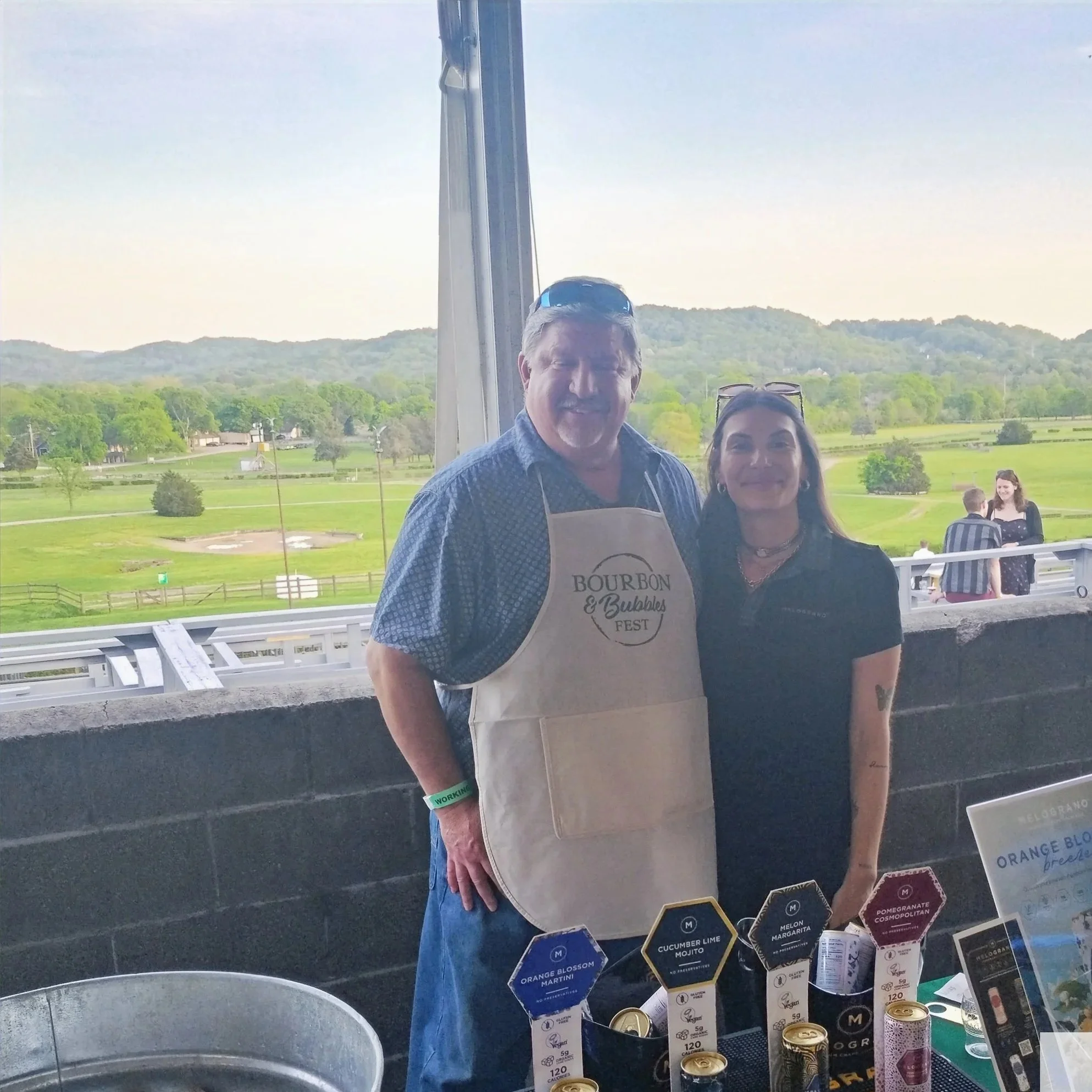 Two people standing near a beverage stand outdoors with green hills behind them. The man is wearing a bourbon and bubbles fest apron.