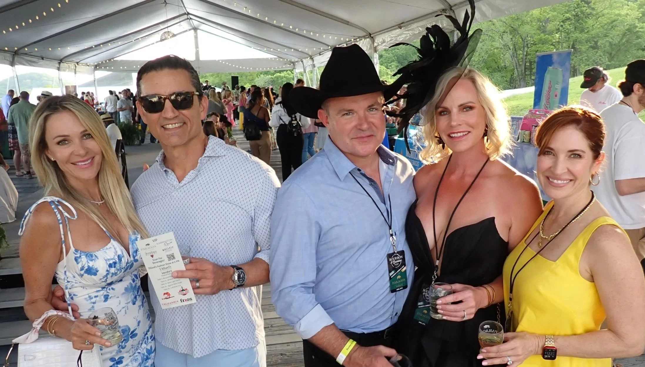 Group of five adults at an outdoor event under a large tent, with two women on each end and two men in the middle. They are smiling and holding drinks, dressed in summery attire, with some wearing accessories like hats and sunglasses. Background show