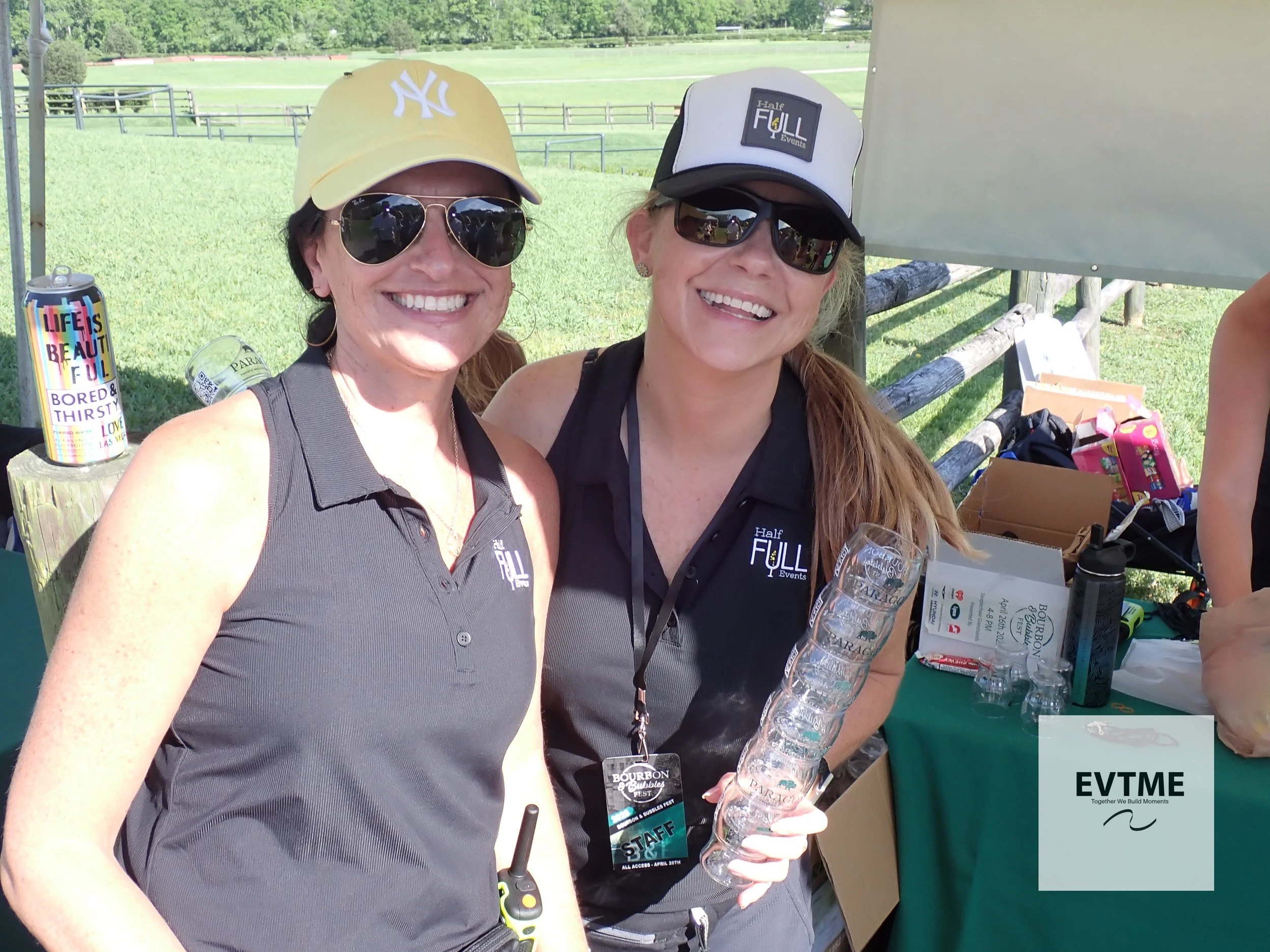 Two women smiling at an outdoor event, wearing black shirts with 'Half Full Events' logo, sunglasses, and caps; one holds an empty glass in hand, with a table of supplies and a water bottle nearby.