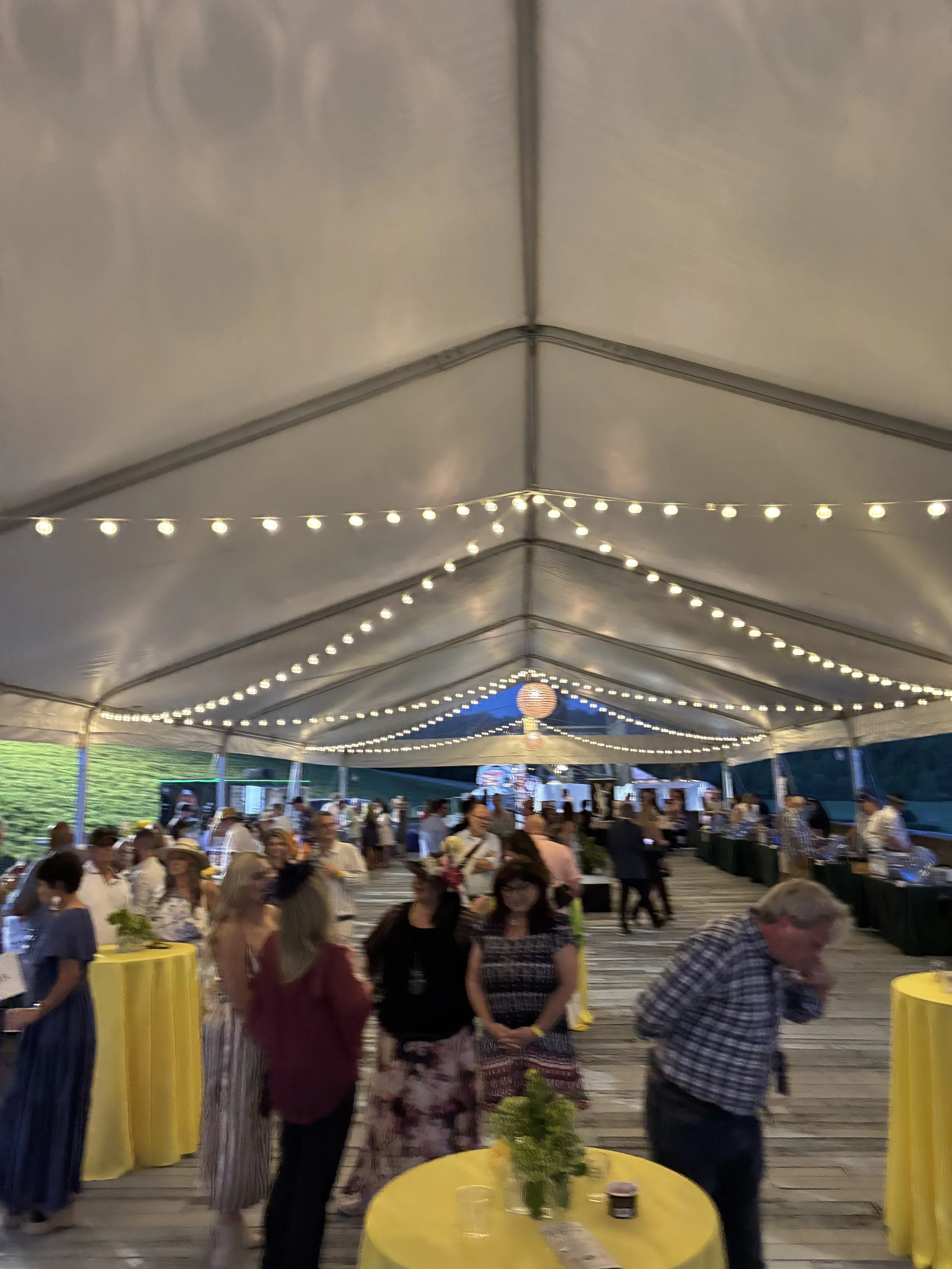 People gathered at a celebratory event under a large tent decorated with string lights. There are high tables with yellow tablecloths, some with floral centerpieces, and a buffet or serving area visible in the background.