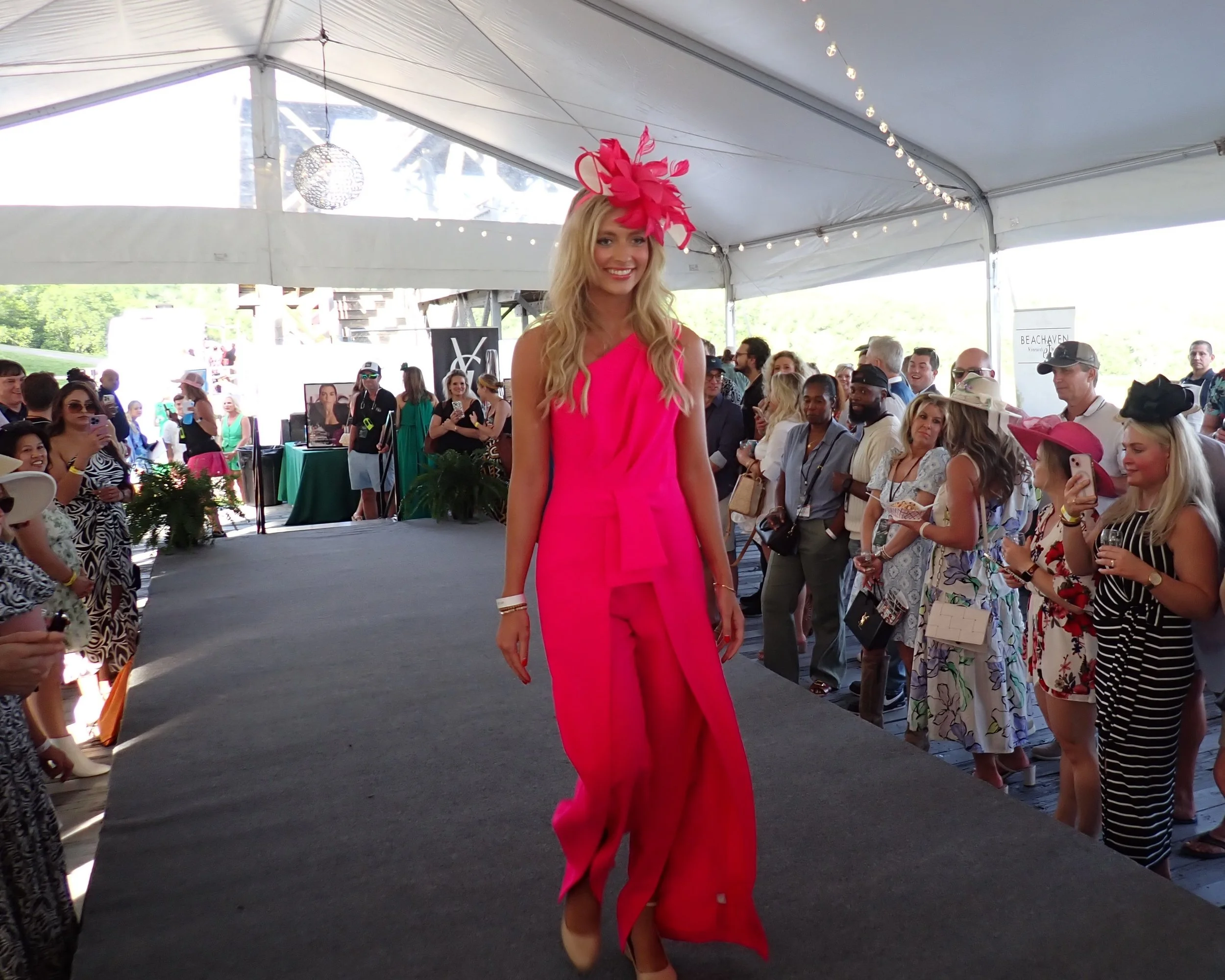 A woman walking on a runway wearing a bright pink outfit and a matching pink fascinator at an outdoor fashion event with an audience and photographers watching.