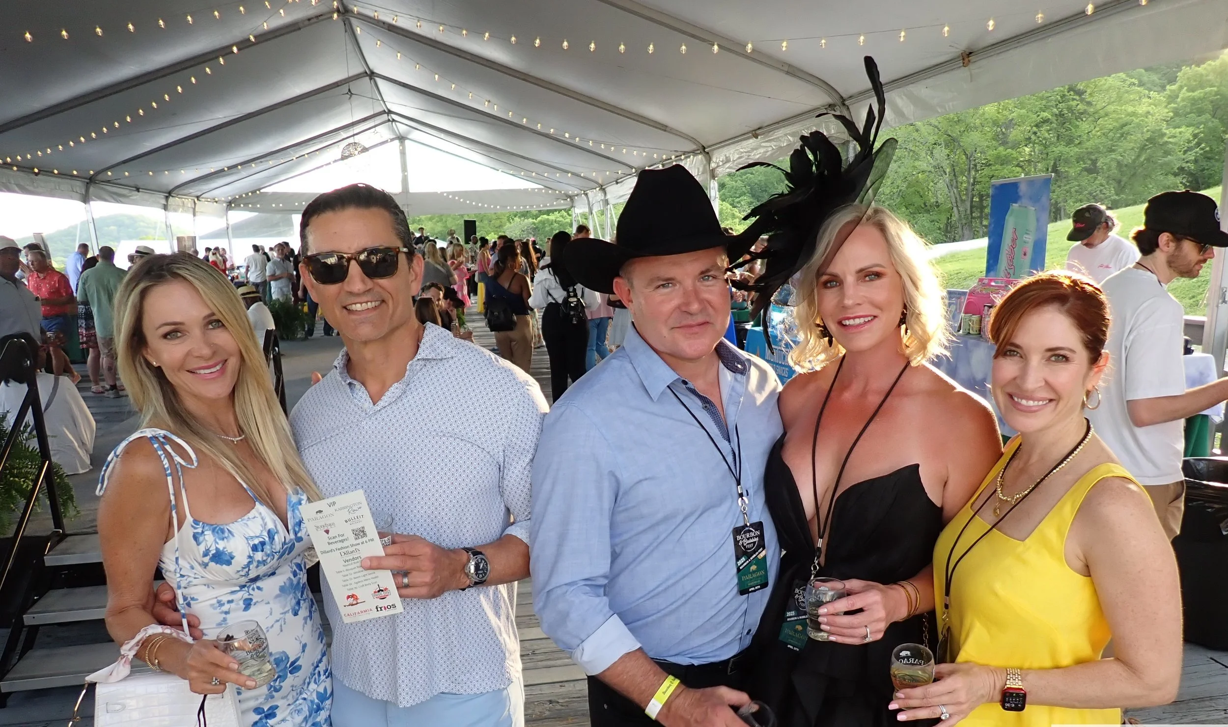Group of five people smiling at a social event inside a large tent with string lights, with others in the background outdoors on a bright day.