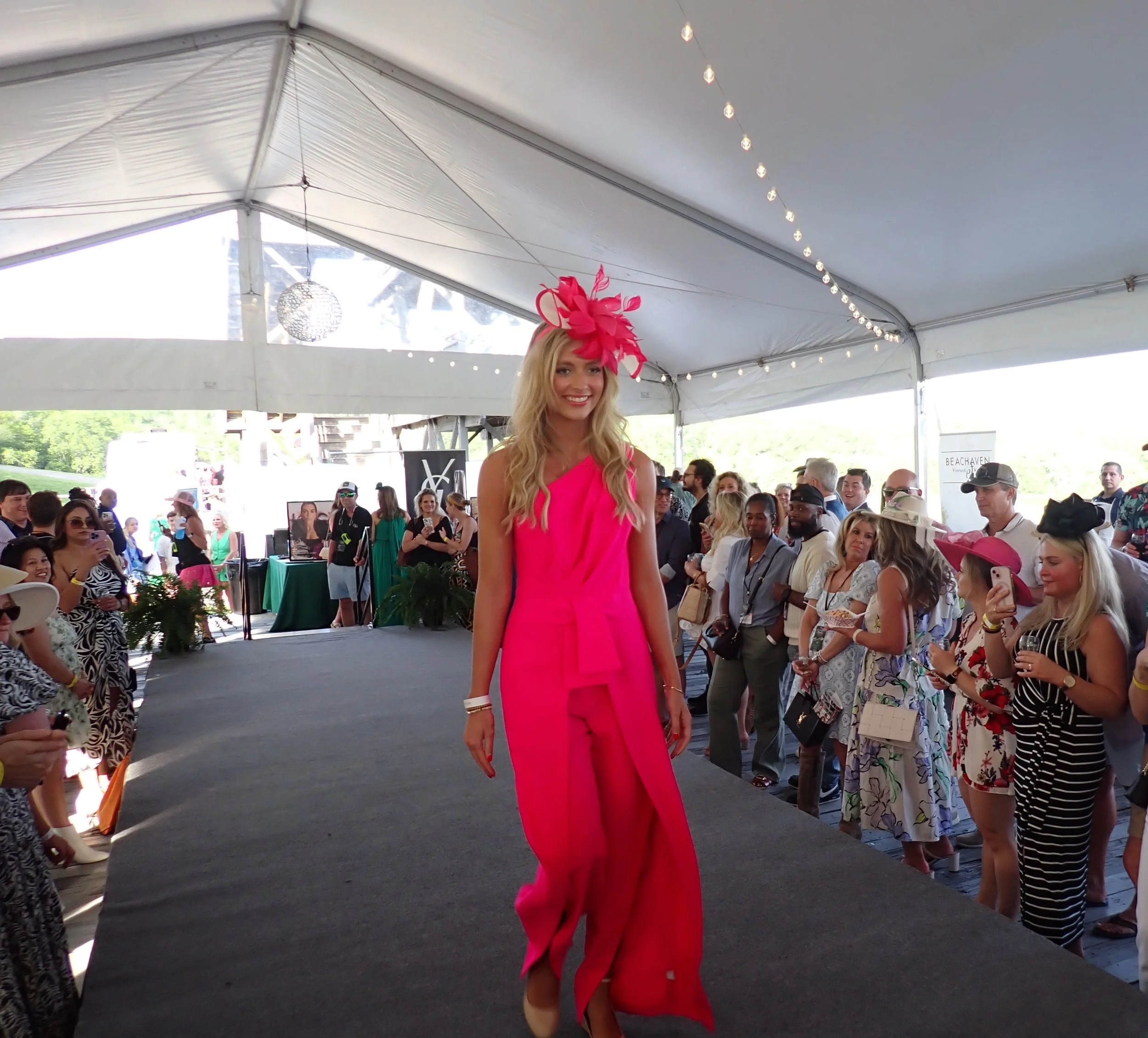 A woman in a bright pink jumpsuit with a matching hat featuring pink feathers walks on a runway at a fashion event, with an audience of mostly women, some taking photos, in a large white tent.